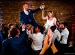 Hora dance with bride and groom on chairs – The bride and groom are lifted high on chairs during the lively Hora dance, laughing and holding hands as guests cheer.