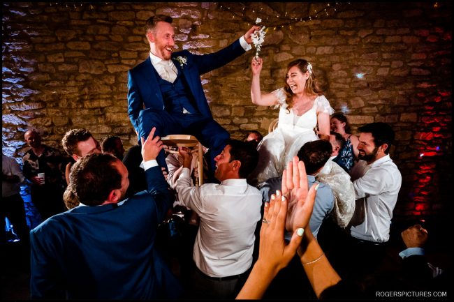 Outdoor Chuppah at Brympton House 083 Hora dance with bride and groom on chairs – The bride and groom are lifted high on chairs during the lively Hora dance, laughing and holding hands as guests cheer.