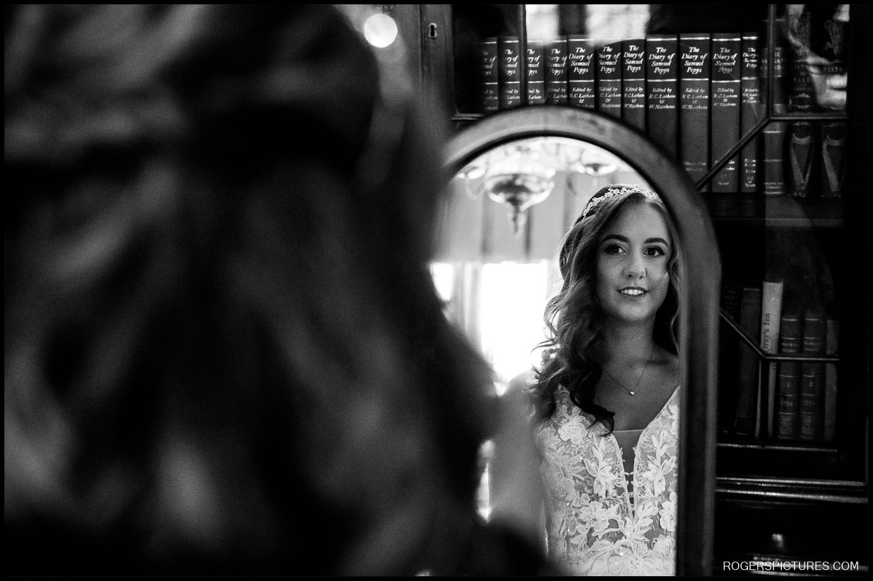 Bride’s Reflection in Mirror – A beautiful black and white portrait of the bride smiling at her reflection, framed by vintage books in the background.