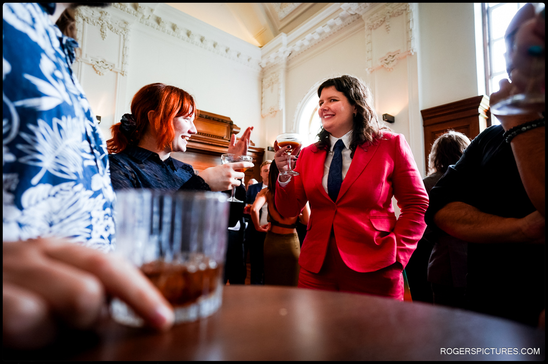 A wedding guest in a bold red suit shares a lively conversation over cocktails in an elegant venue.