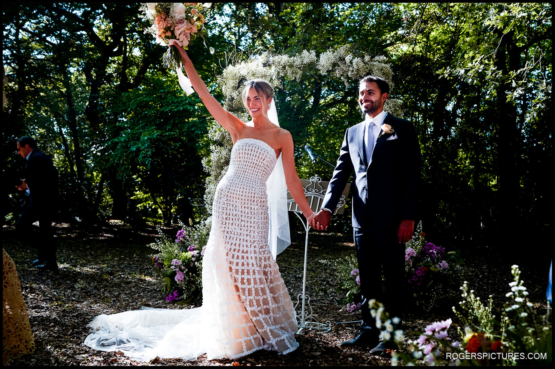 Bride raises her bouquet in celebration while holding hands with the groom after their outdoor ceremony in Tuscany.