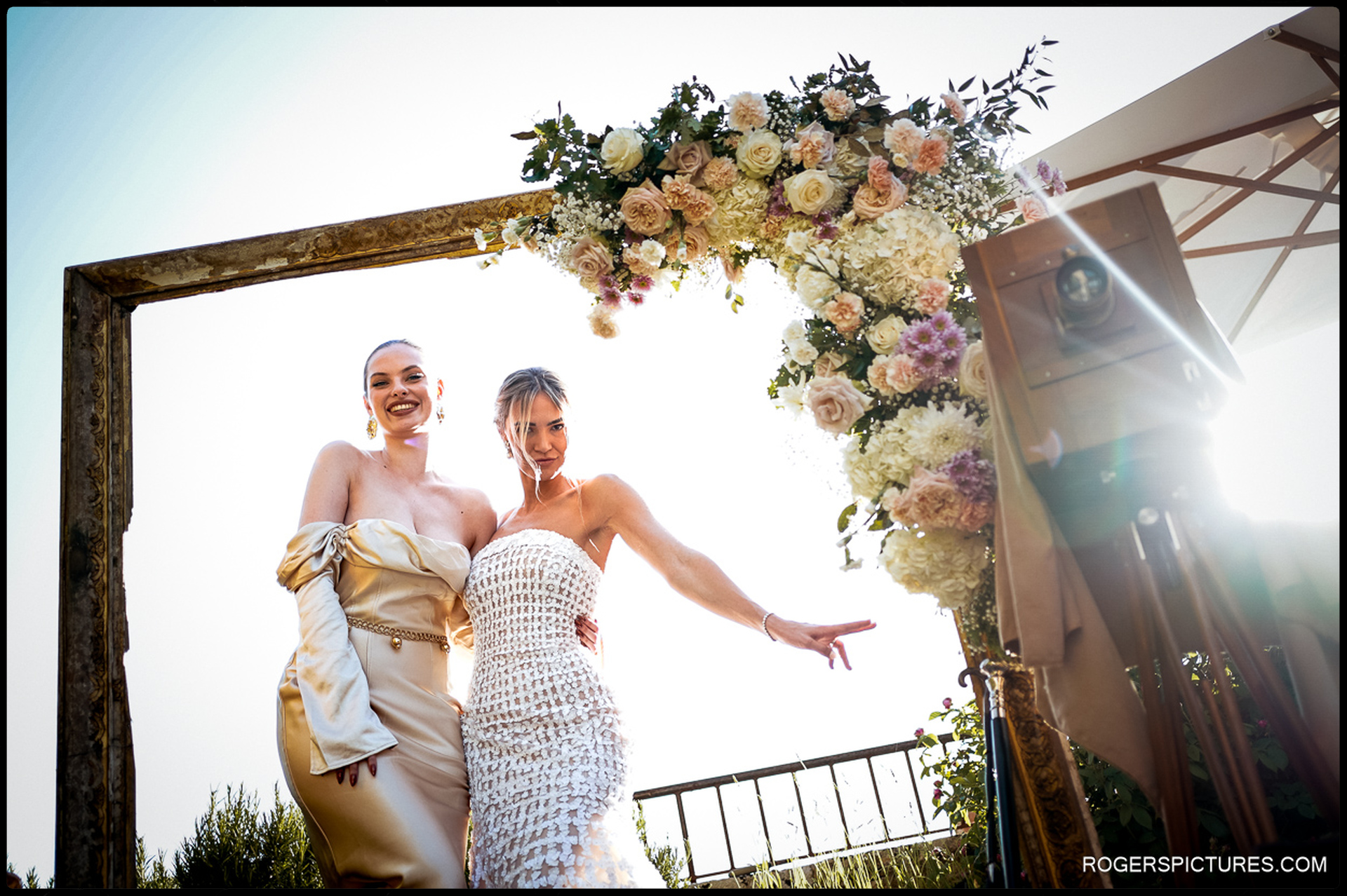Bride and bridesmaid pose together under a floral arch at a Tuscan wedding celebration.