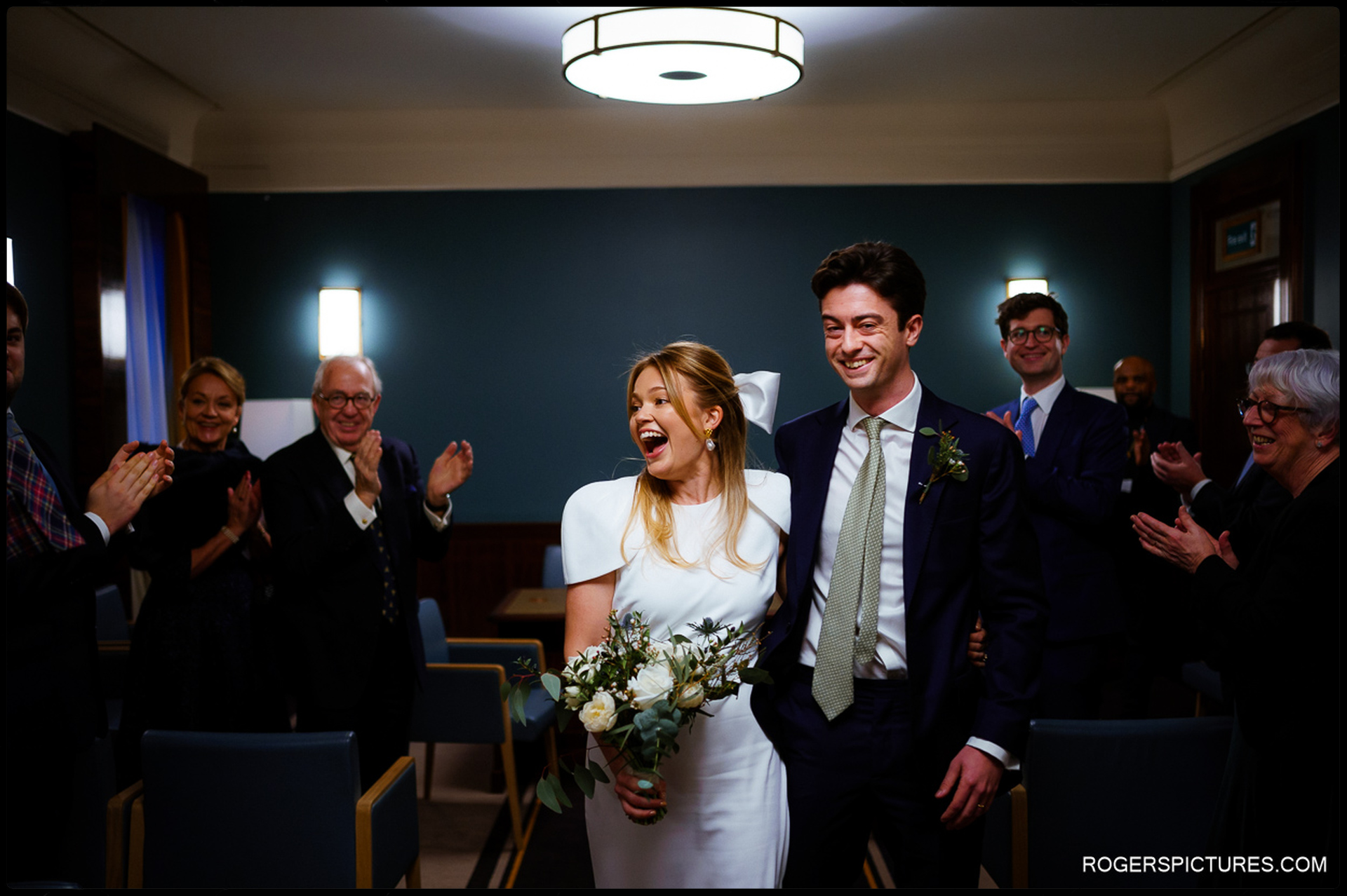 Bride and groom walk back up the aisle, smiling as guests applaud their Short Wedding at Hackney Town Hall.
