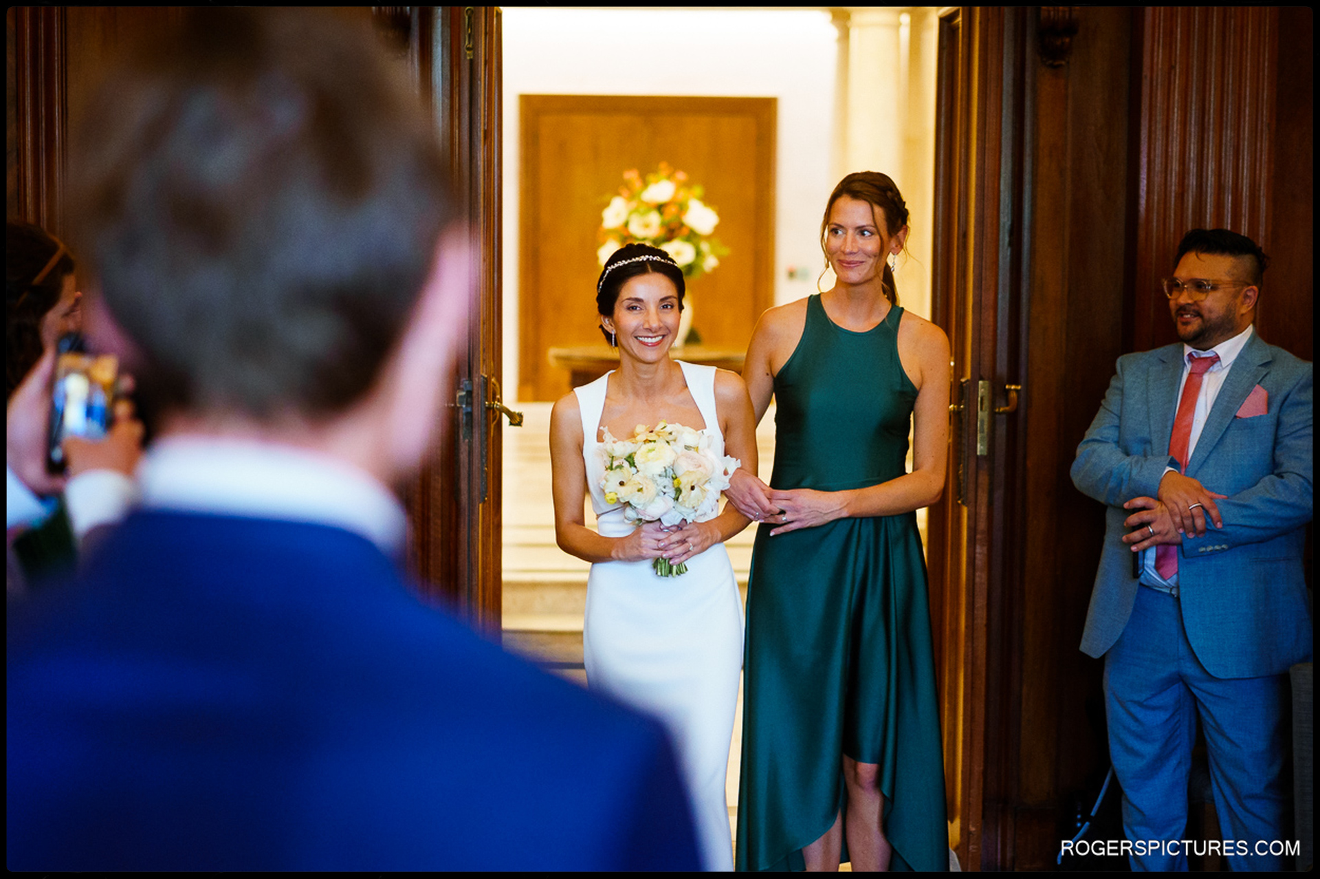 Bride and bridesmaid entering the ceremony room at Marylebone Town Hall, greeted by guests.