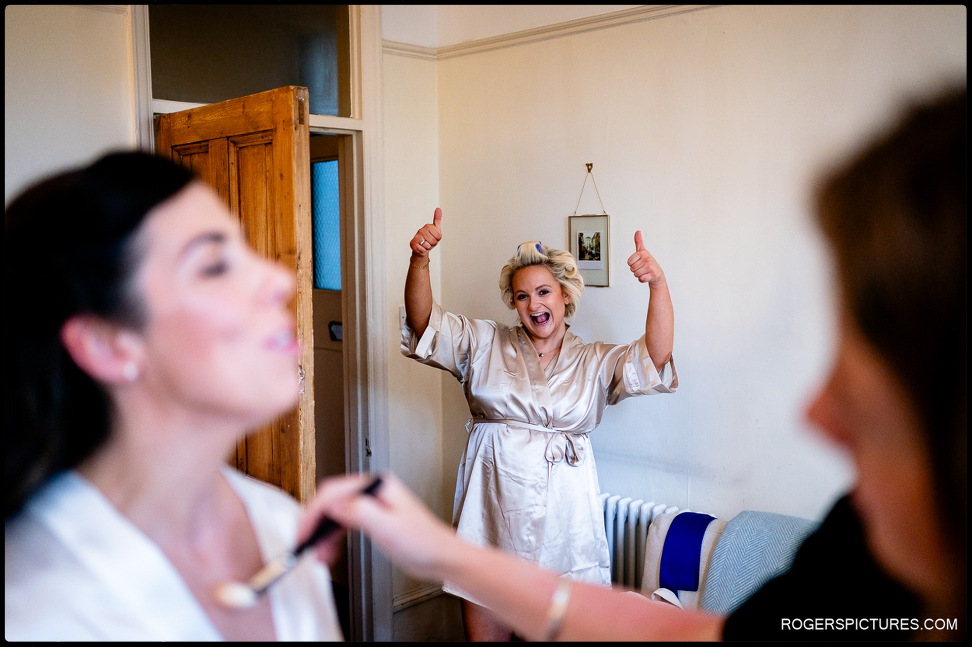 Bride getting makeup done as bridesmaid gives thumbs up in background.
