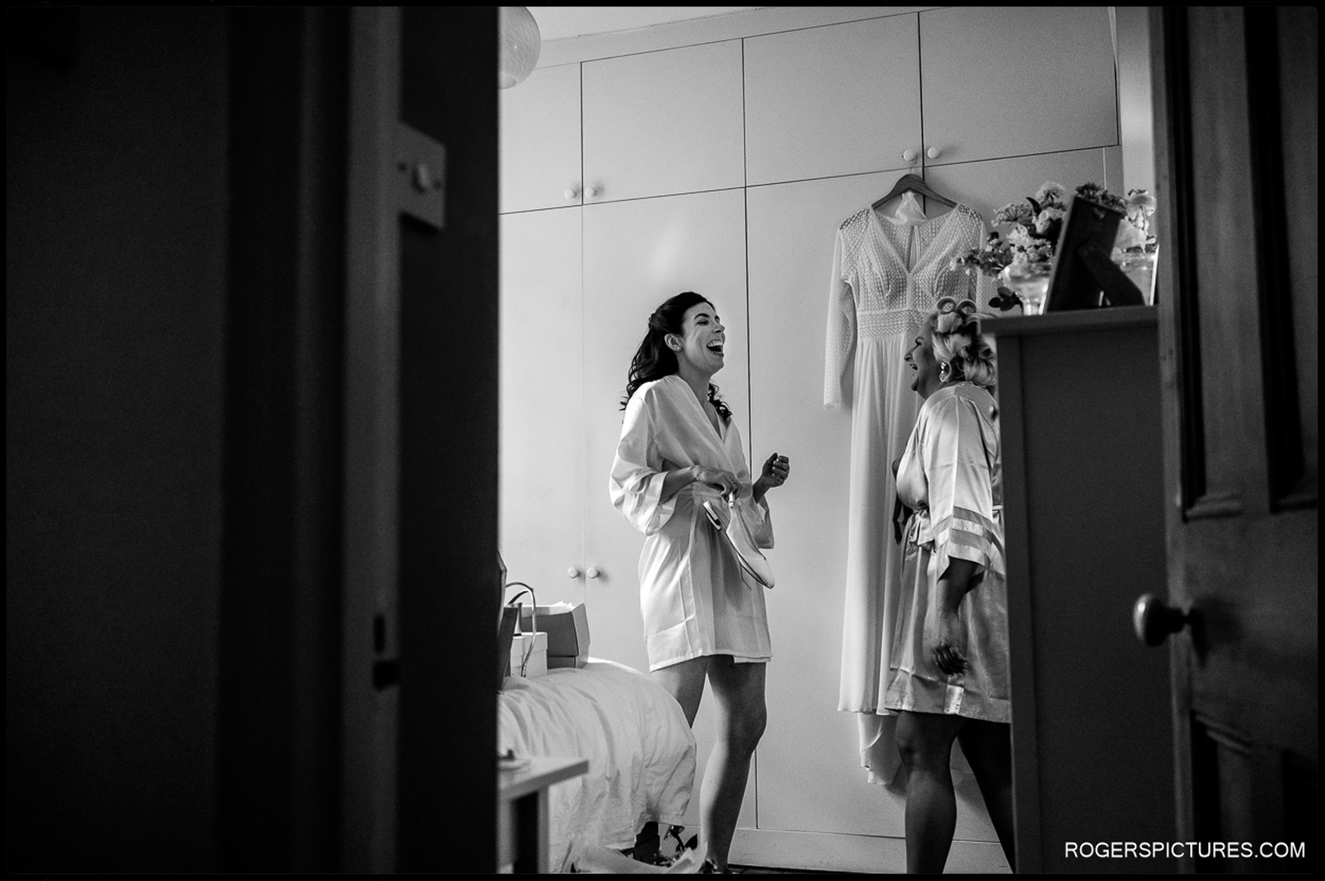 Bride laughing with bridesmaid while getting ready next to the wedding dress.