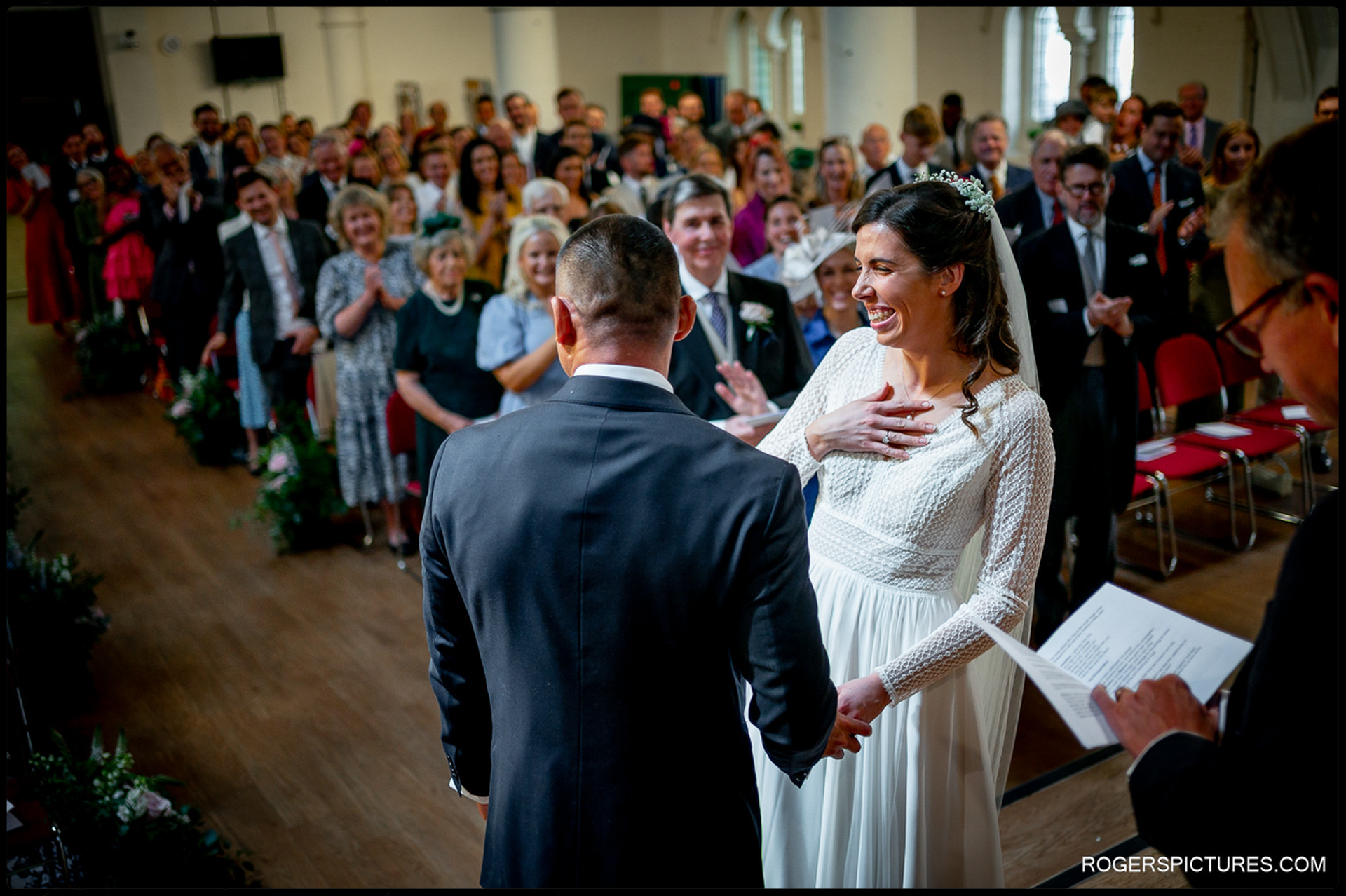 Bride laughing and holding the groom’s hand during vows, surrounded by smiling guests.