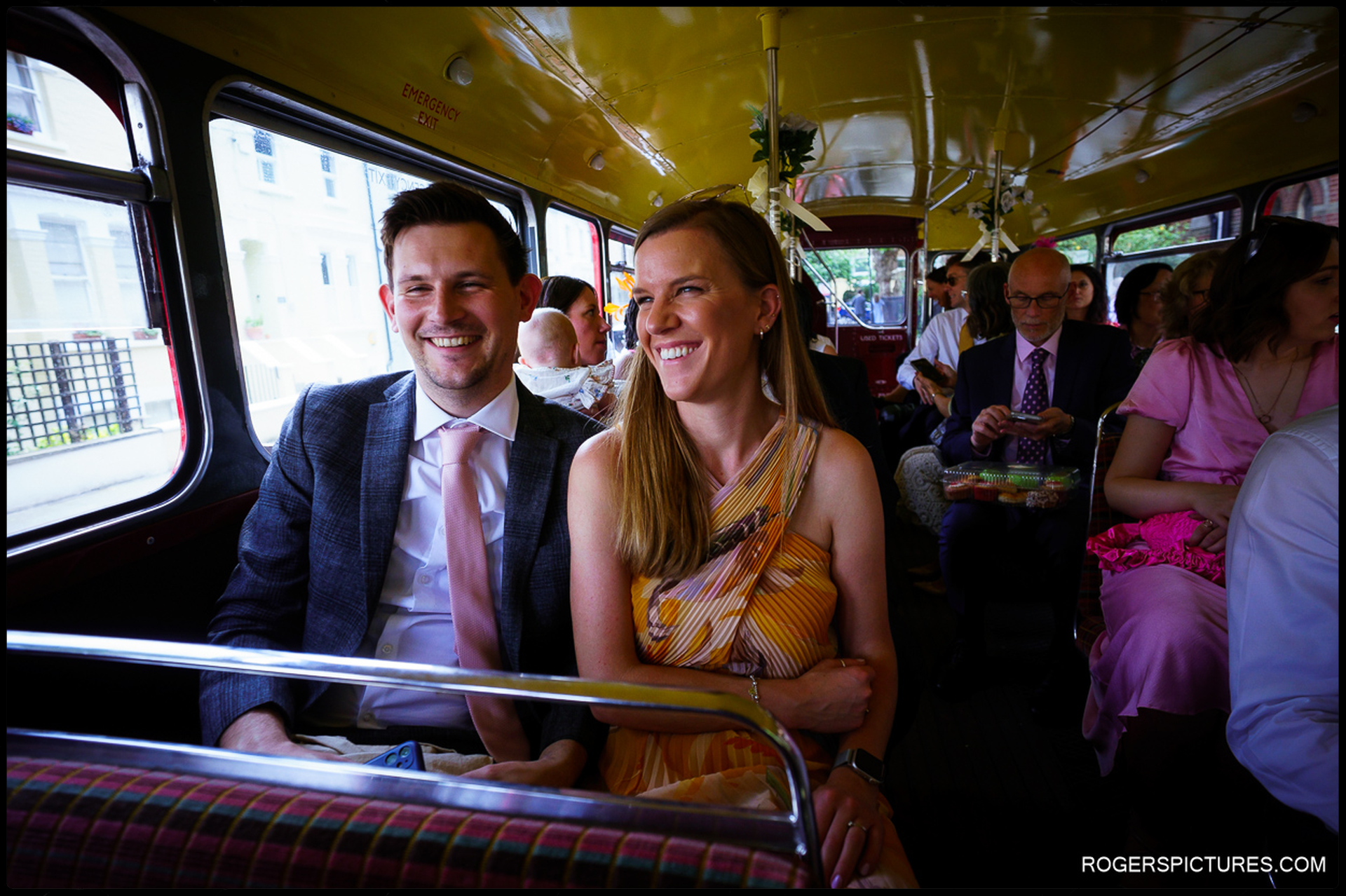 Wedding guests smiling on board a red double-decker bus en route to the reception.
