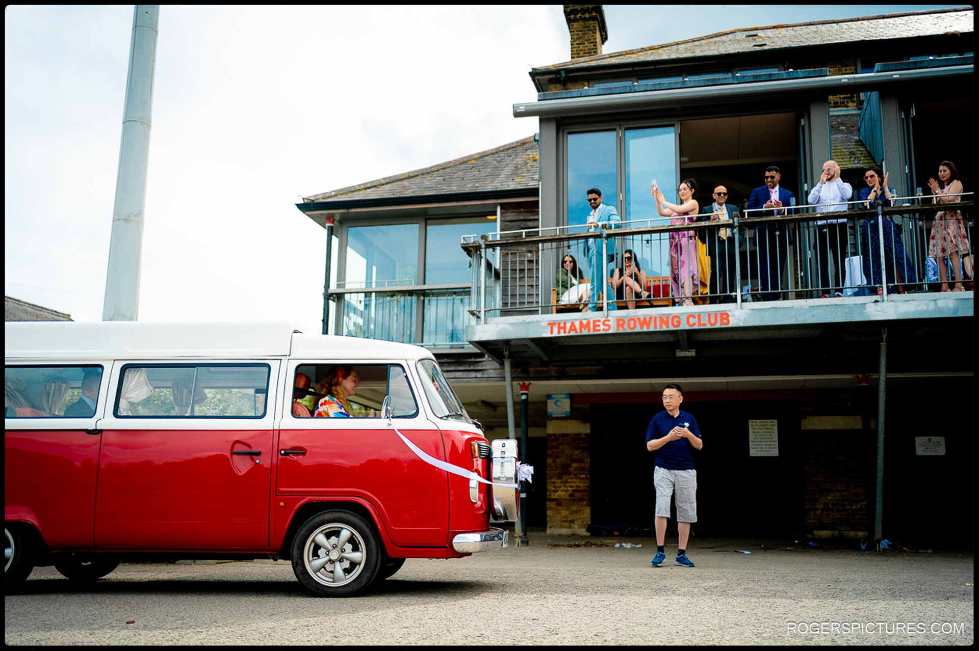 Red vintage camper van arriving at Thames Rowing Club while guests cheer from the balcony.