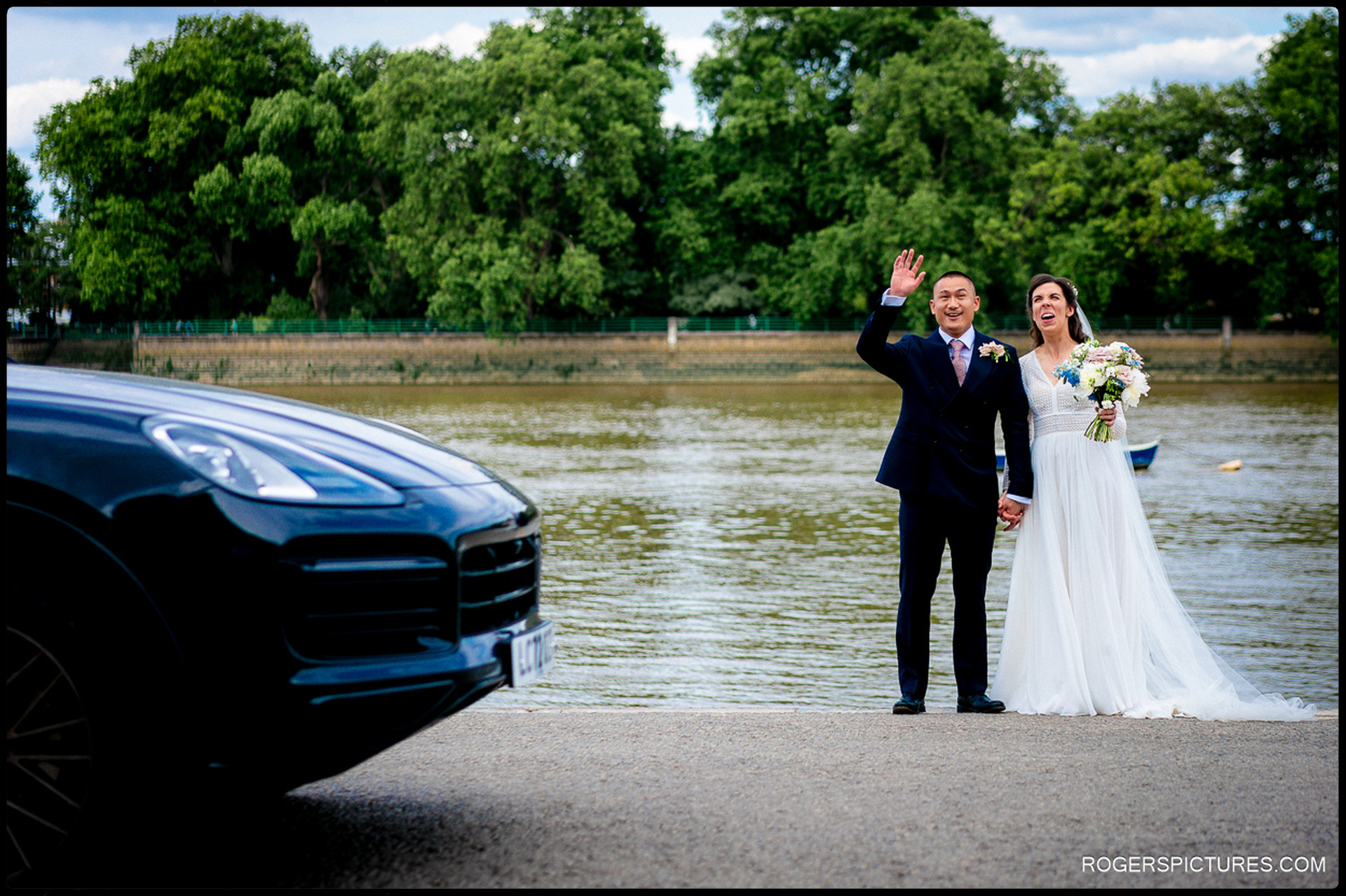 Bride and groom waving and laughing beside the Thames River, with car in foreground.