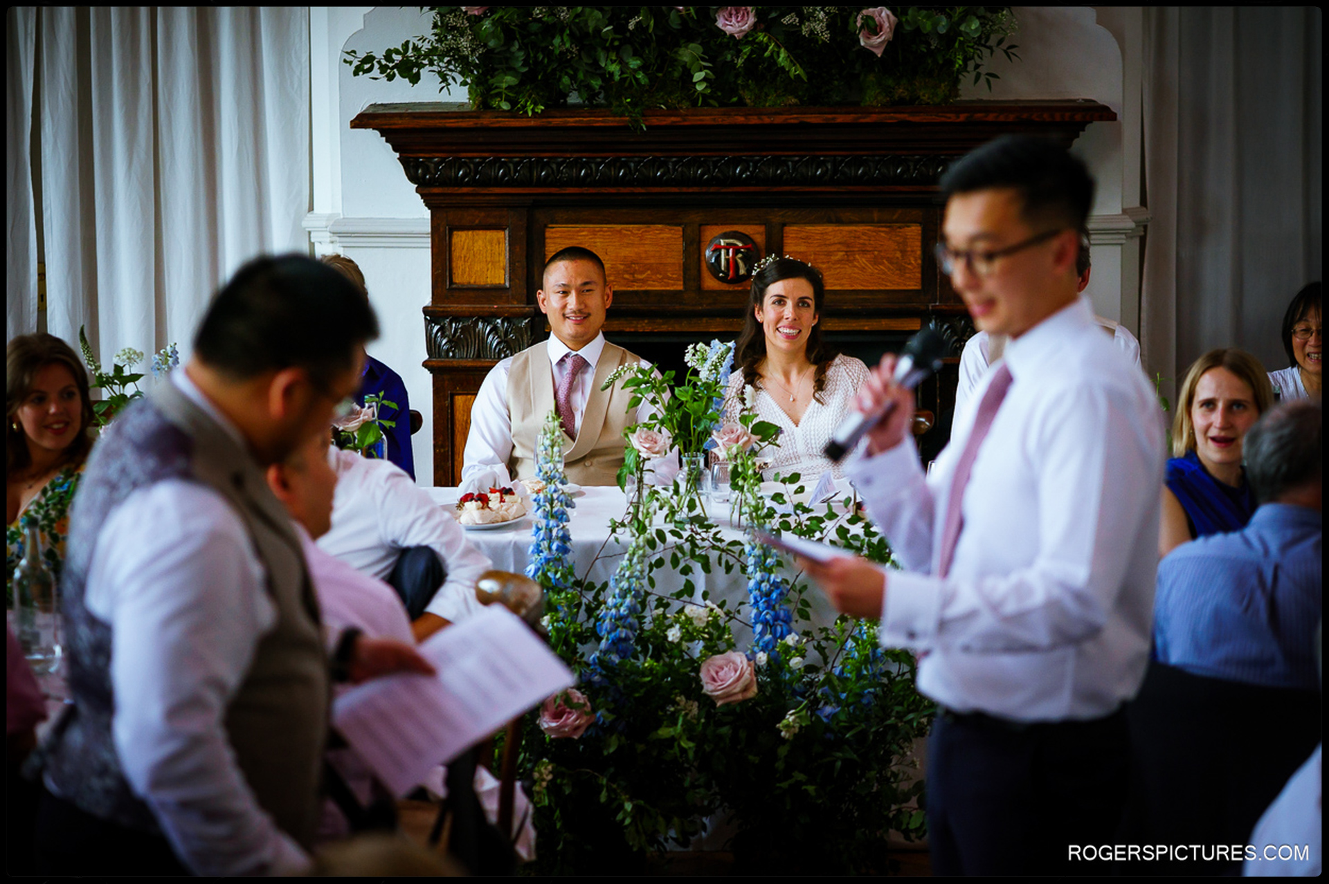 Bride and groom smiling at the top table while listening to a speech, with floral arrangements in front of them.