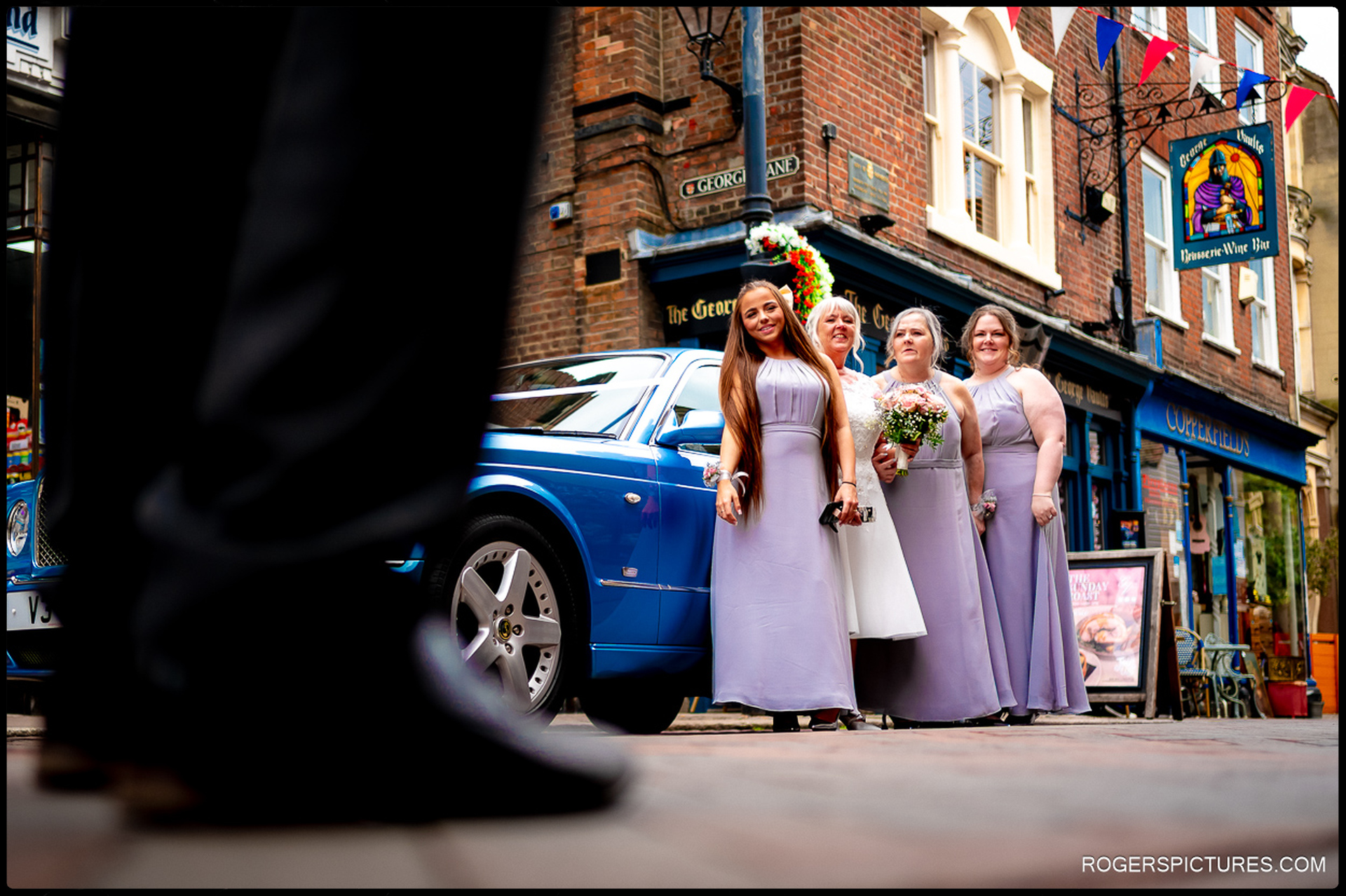 Bridesmaids in lilac dresses and bride pose beside a blue Bentley on George Lane in Rochester, captured from behind a guest’s silhouette.
