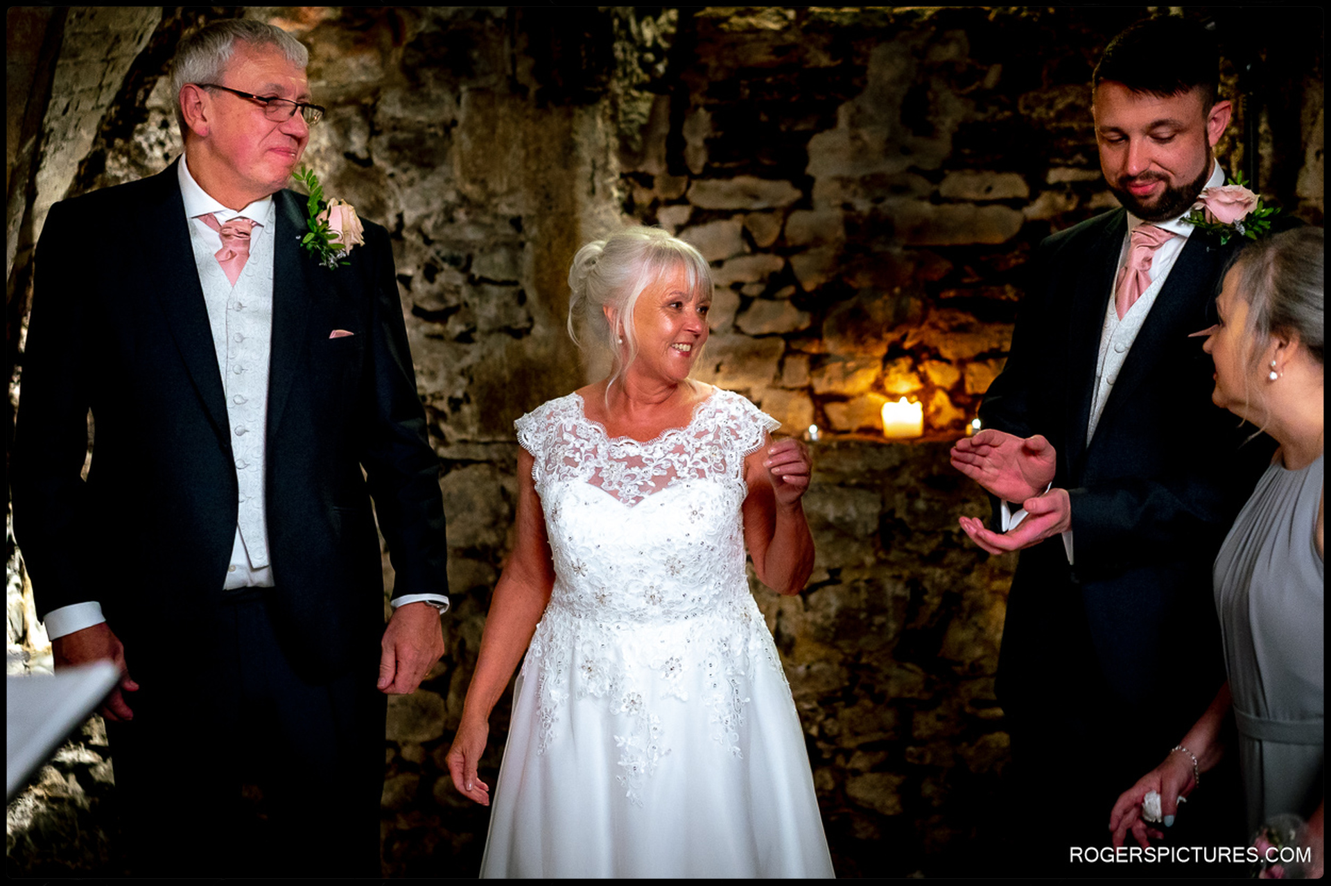 Bride in lace dress smiles during the ceremony, flanked by groom and wedding guests against a stone wall lit by candles.