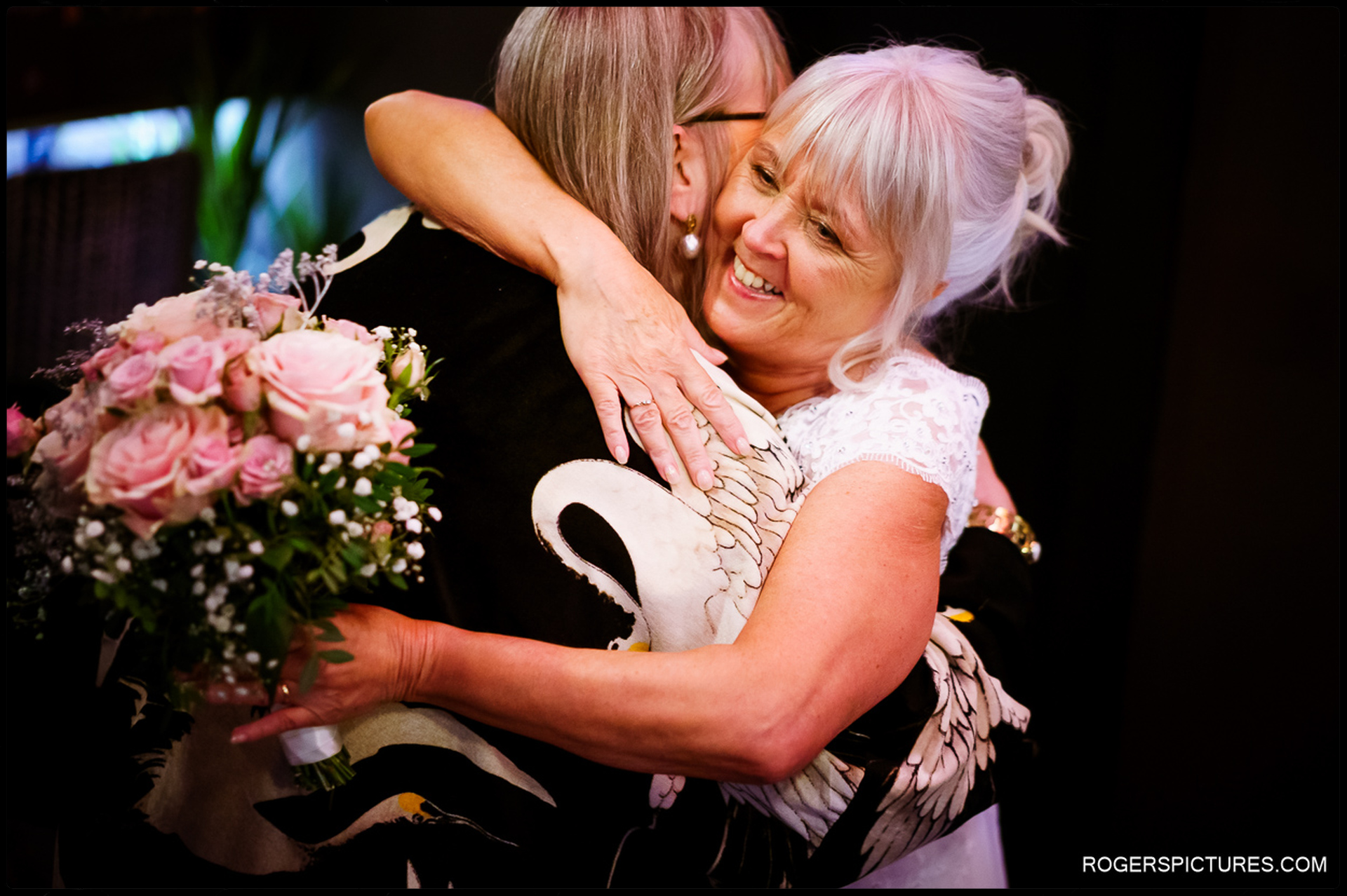 Bride embraces a guest with a joyful smile, holding a bouquet of pink roses.