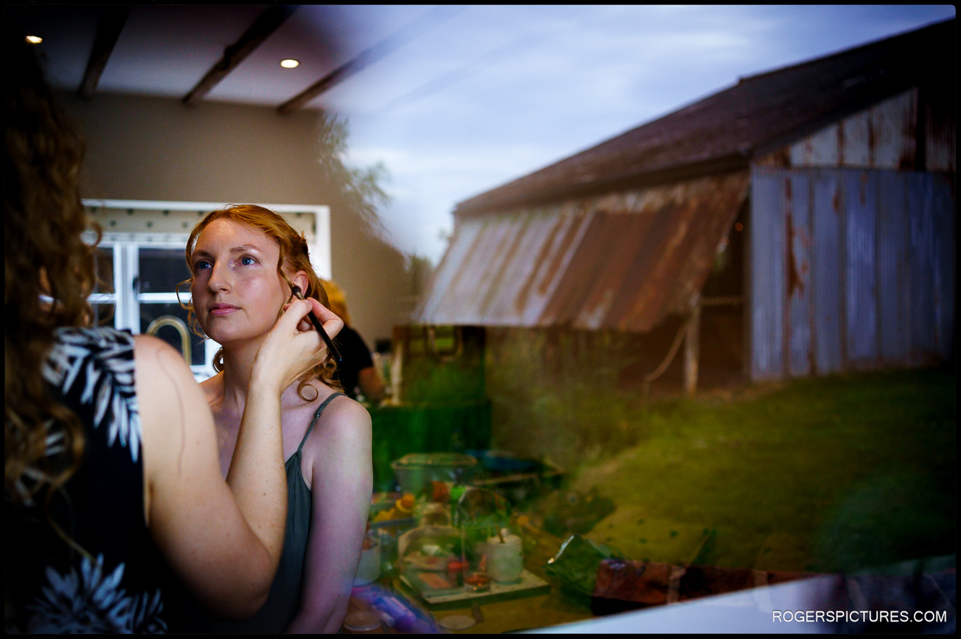 Bride reflected in the window during makeup, with rustic barn at The Firs Hanbury visible outside.