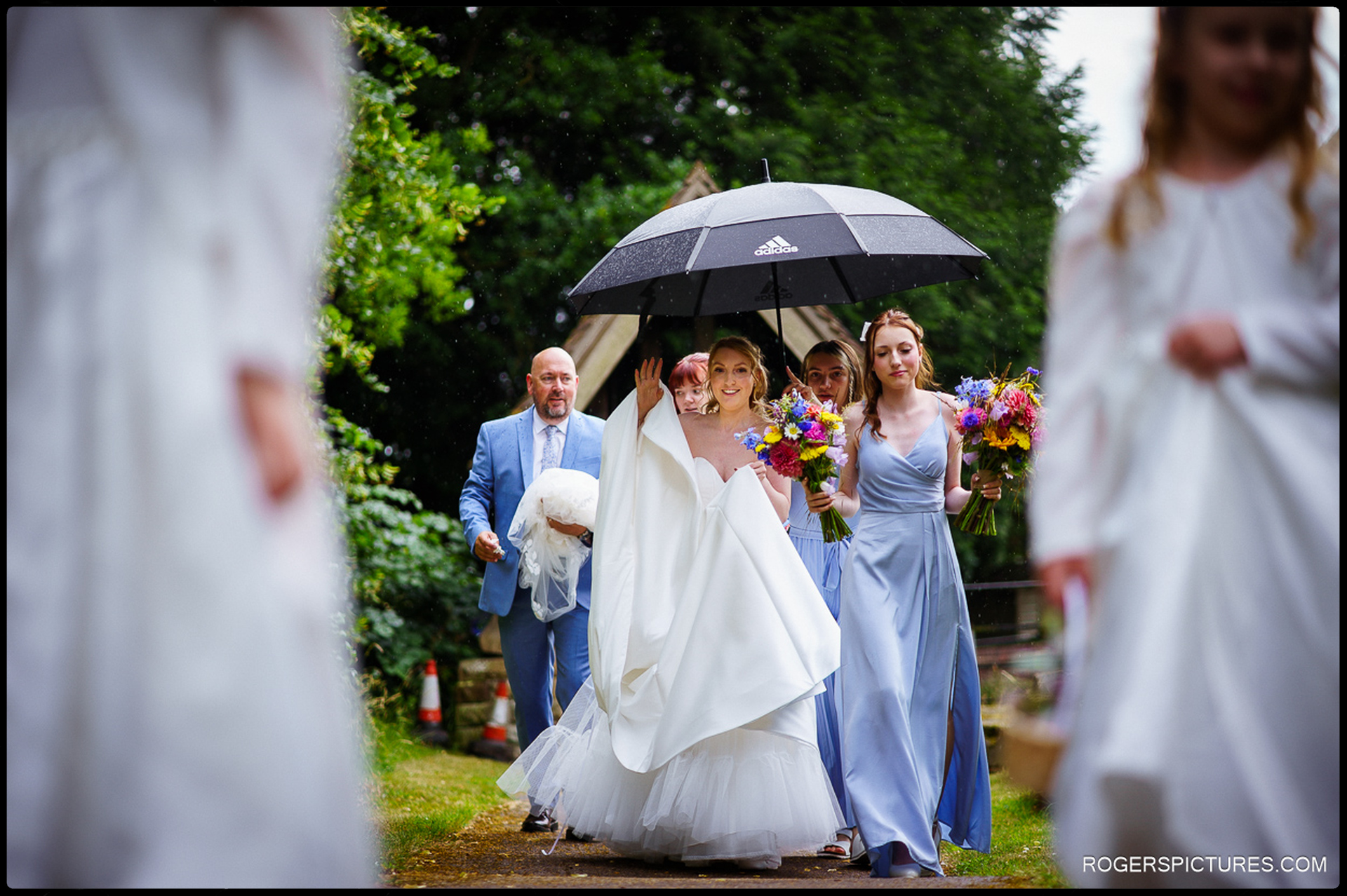 Bride walking to the church under an umbrella with her bridesmaids and father on a rainy June day.