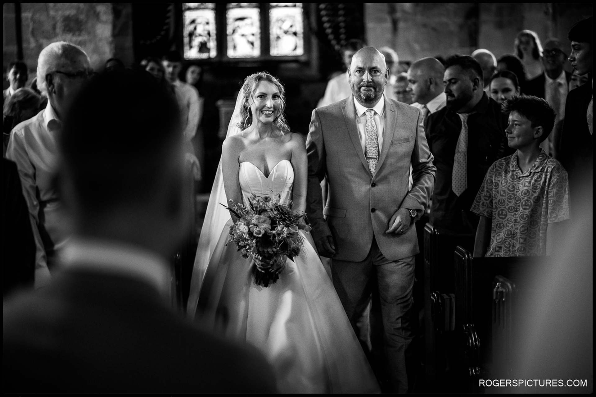Bride and her father walking down the aisle at St John The Baptist Church as guests look on.
