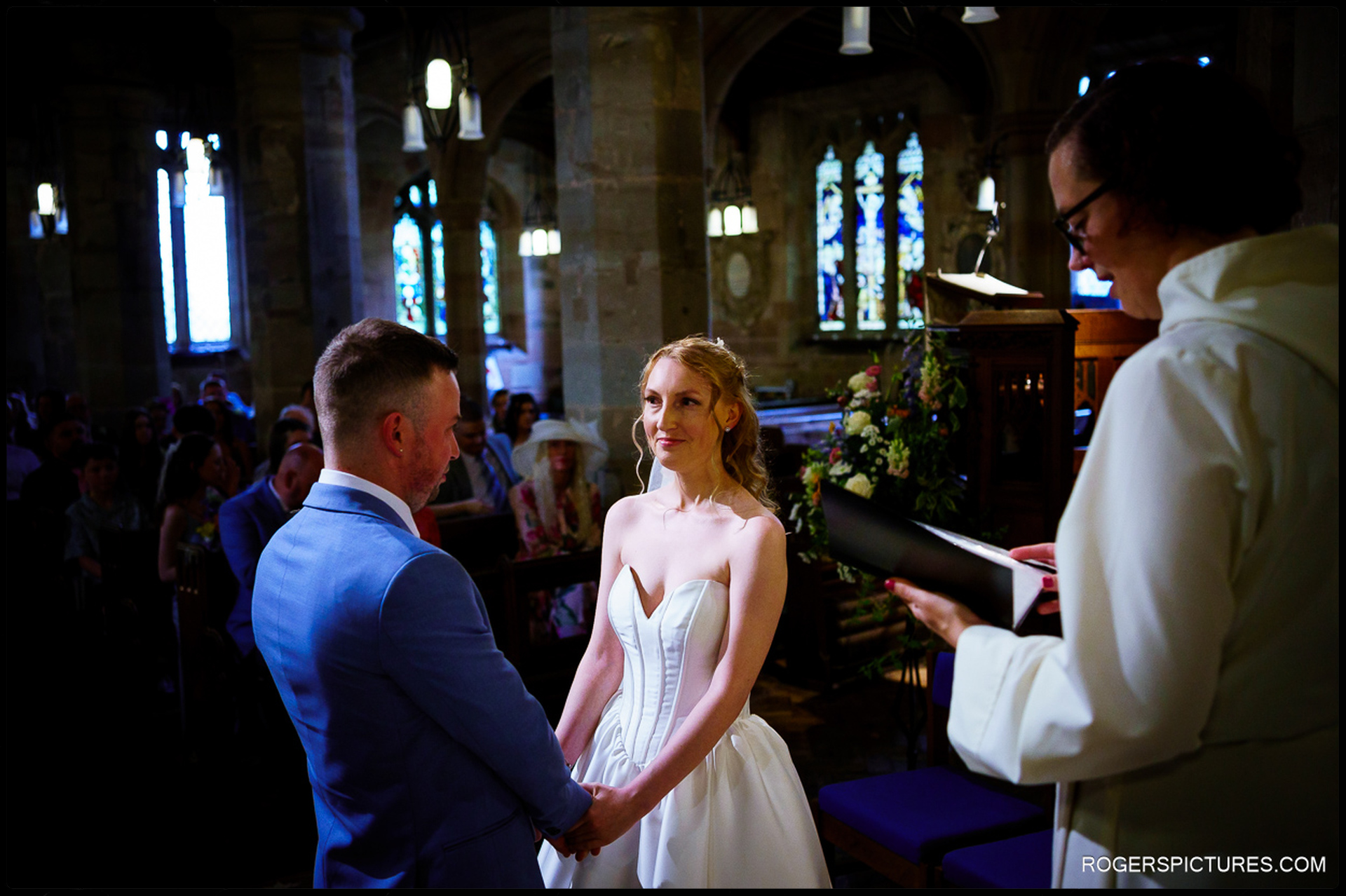 Couple holding hands during the ceremony, with the vicar reading vows at the front of the church.