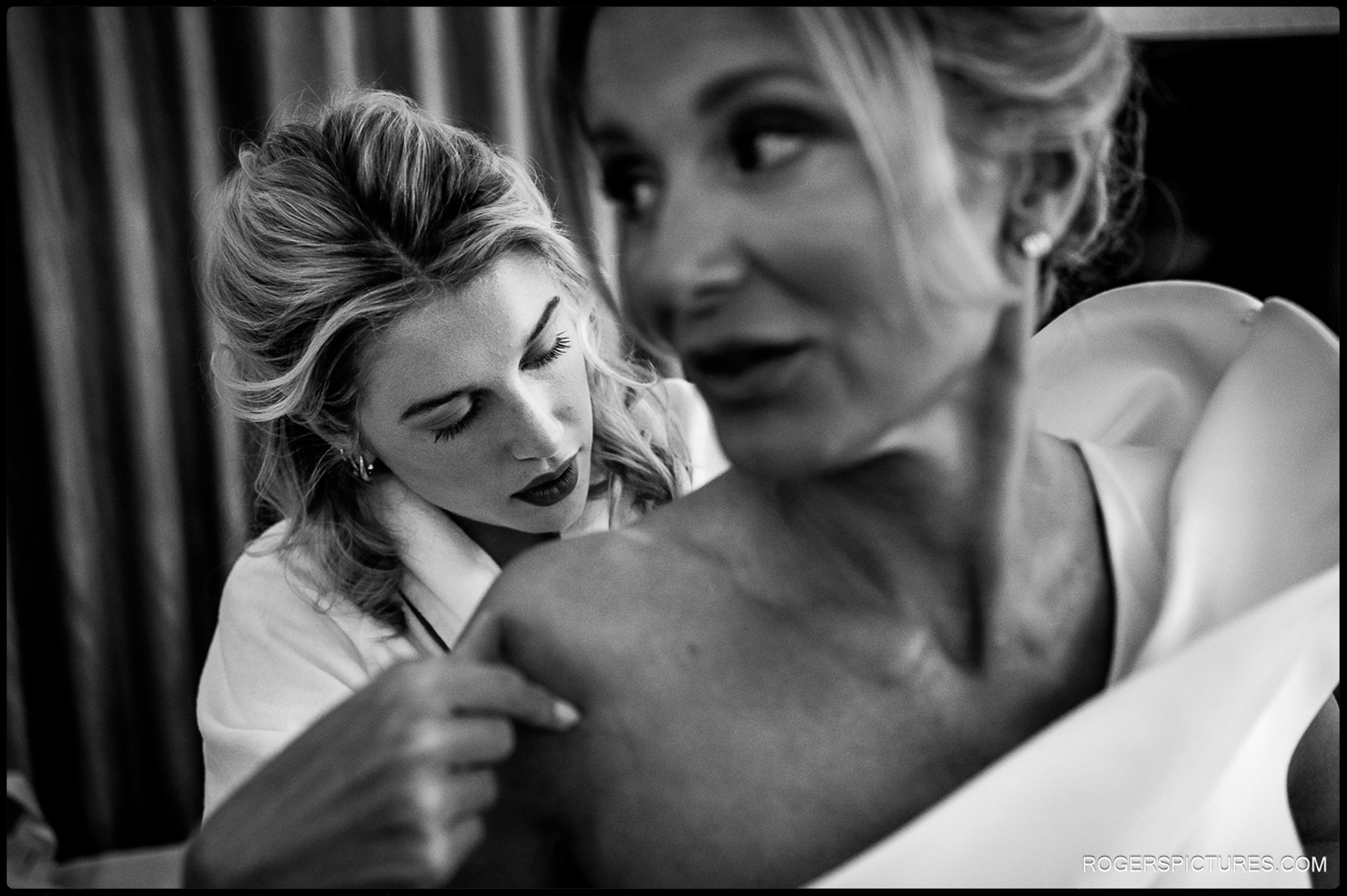 Black and white image of a bridesmaid helping the bride fasten her dress, showing focus and gentle interaction.