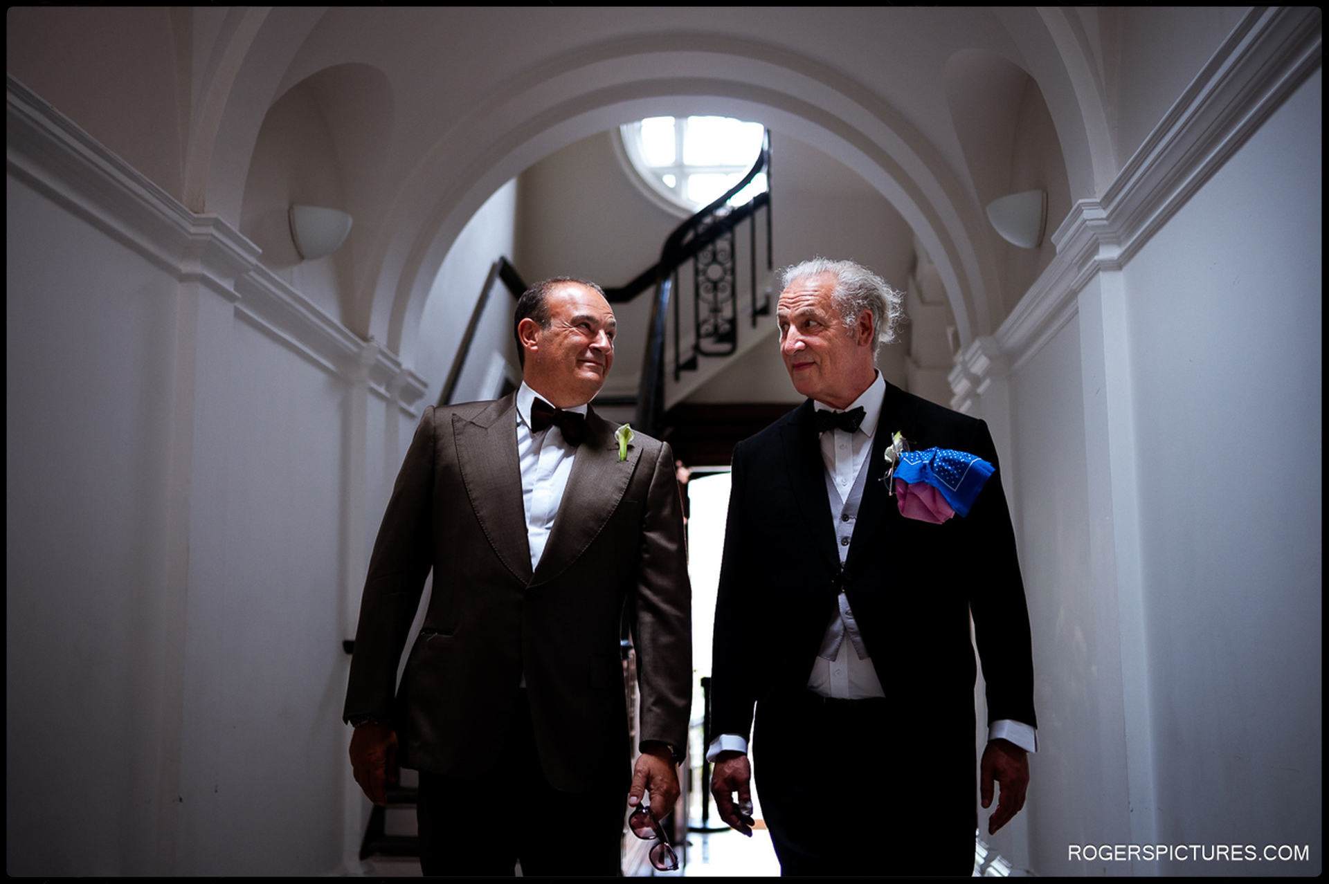 Groom walking through a bright hallway at Chelsea Old Town Hall with a groomsman, both dressed in tuxedos and smiling.