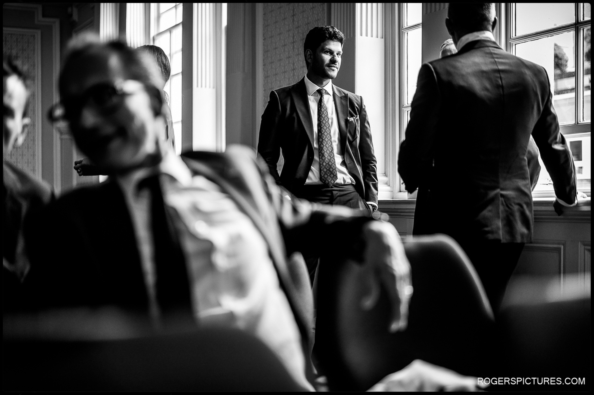 Black and white candid moment of guests chatting before the ceremony at Chelsea Old Town Hall, with natural light streaming through tall windows.