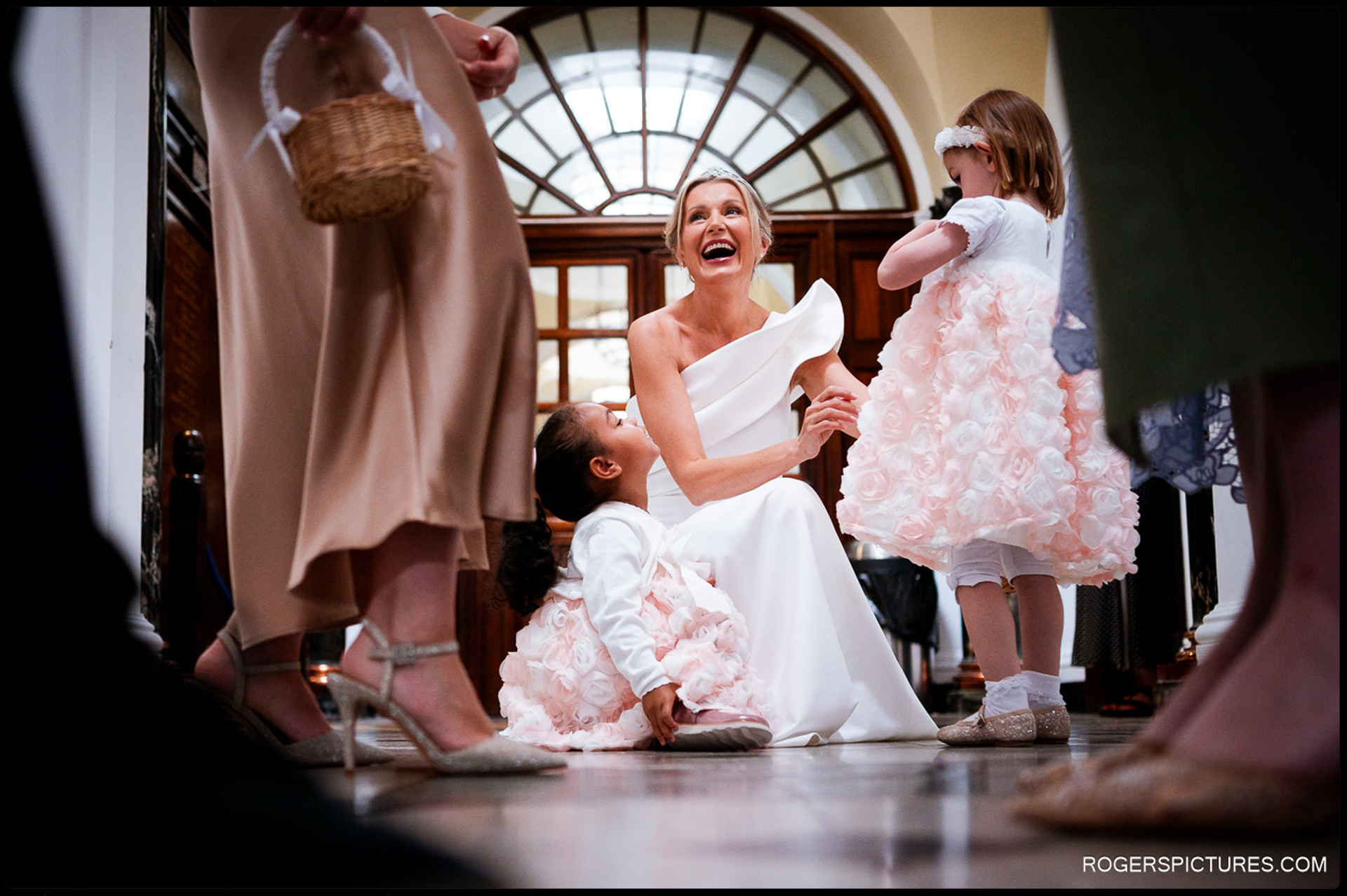 Bride crouching to talk and laugh with young flower girls dressed in pale pink floral dresses at Chelsea Old Town Hall.
