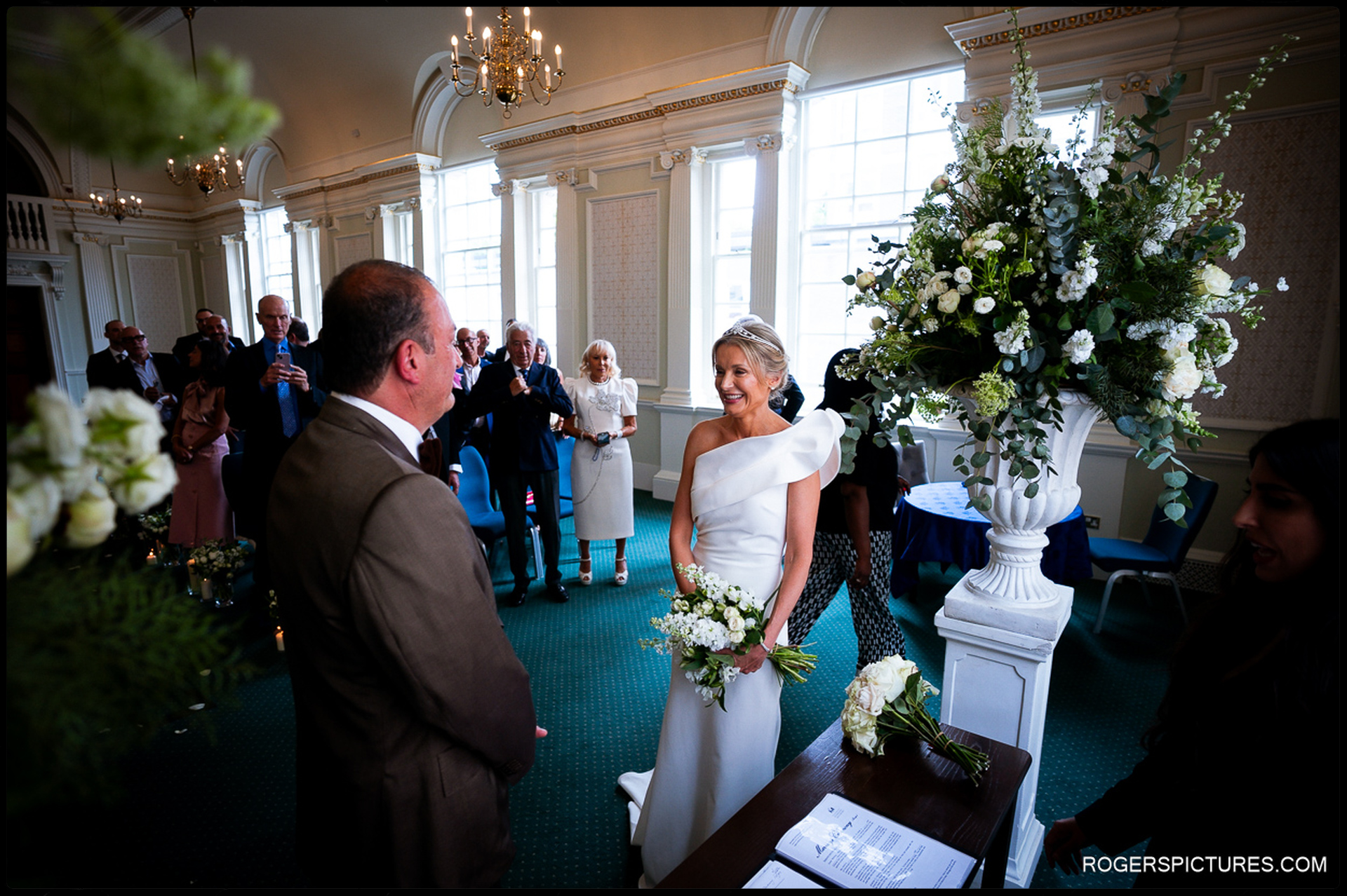 Bride arriving at the top of the aisle, smiling at the groom as guests look on inside the bright, high-ceilinged ceremony room at Chelsea Old Town Hall.