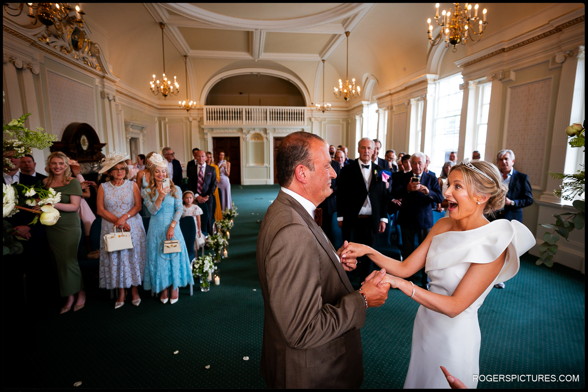 The couple laughing and holding hands mid-ceremony, surrounded by smiling guests inside Chelsea Old Town Hall’s grand room.