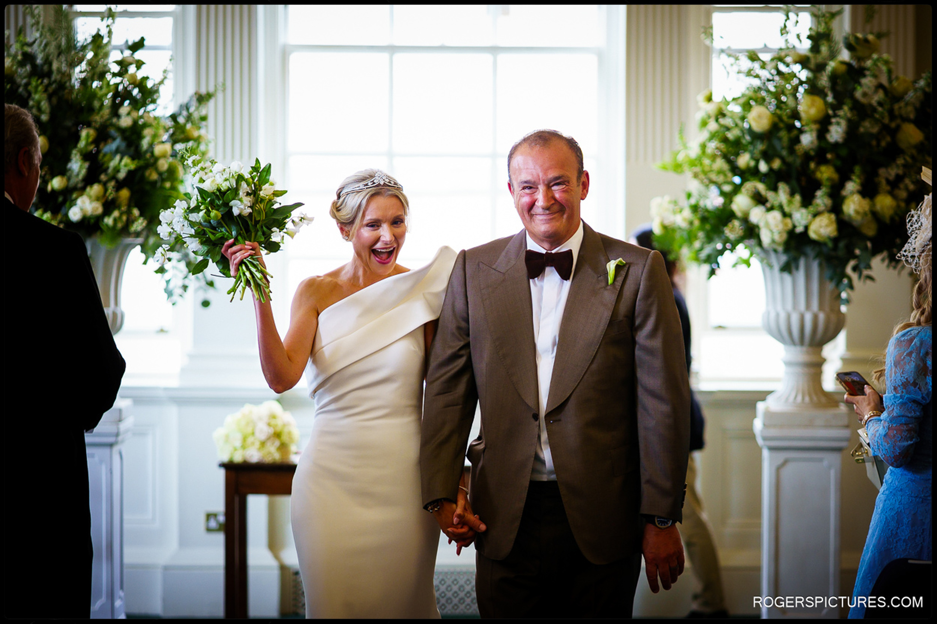 Bride and groom walking back up the aisle, hand in hand, as newlyweds at Chelsea Old Town Hall, with the bride holding her bouquet high in celebration.