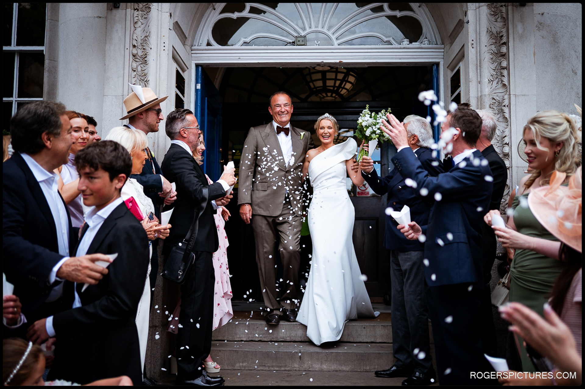 Bride and groom exiting Chelsea Old Town Hall to a joyful confetti throw, surrounded by cheering guests on the steps.
