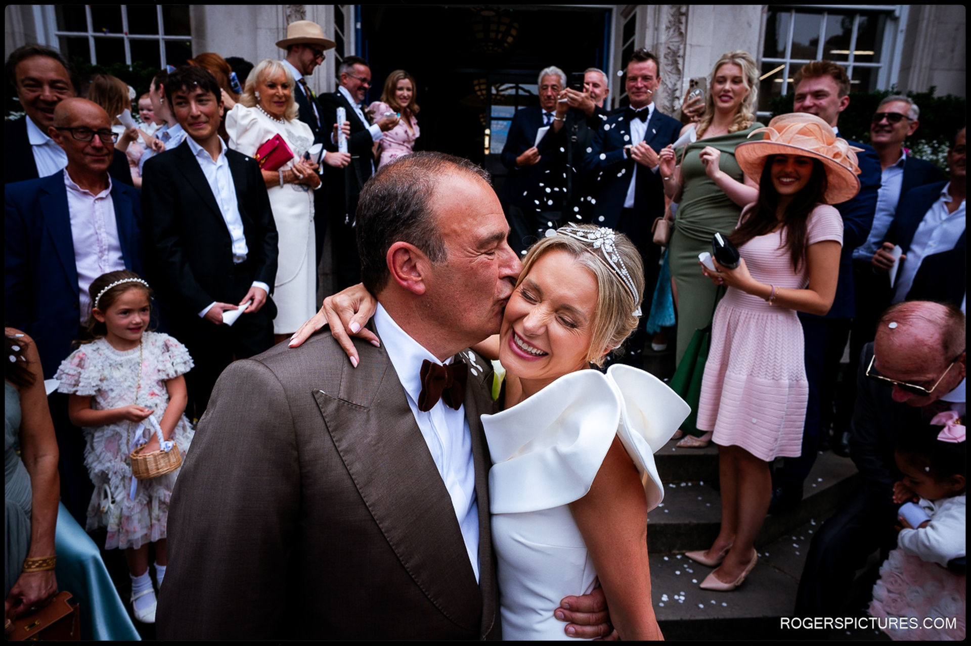 Groom kissing the bride on the cheek outside Chelsea Old Town Hall, surrounded by smiling guests and scattered confetti.