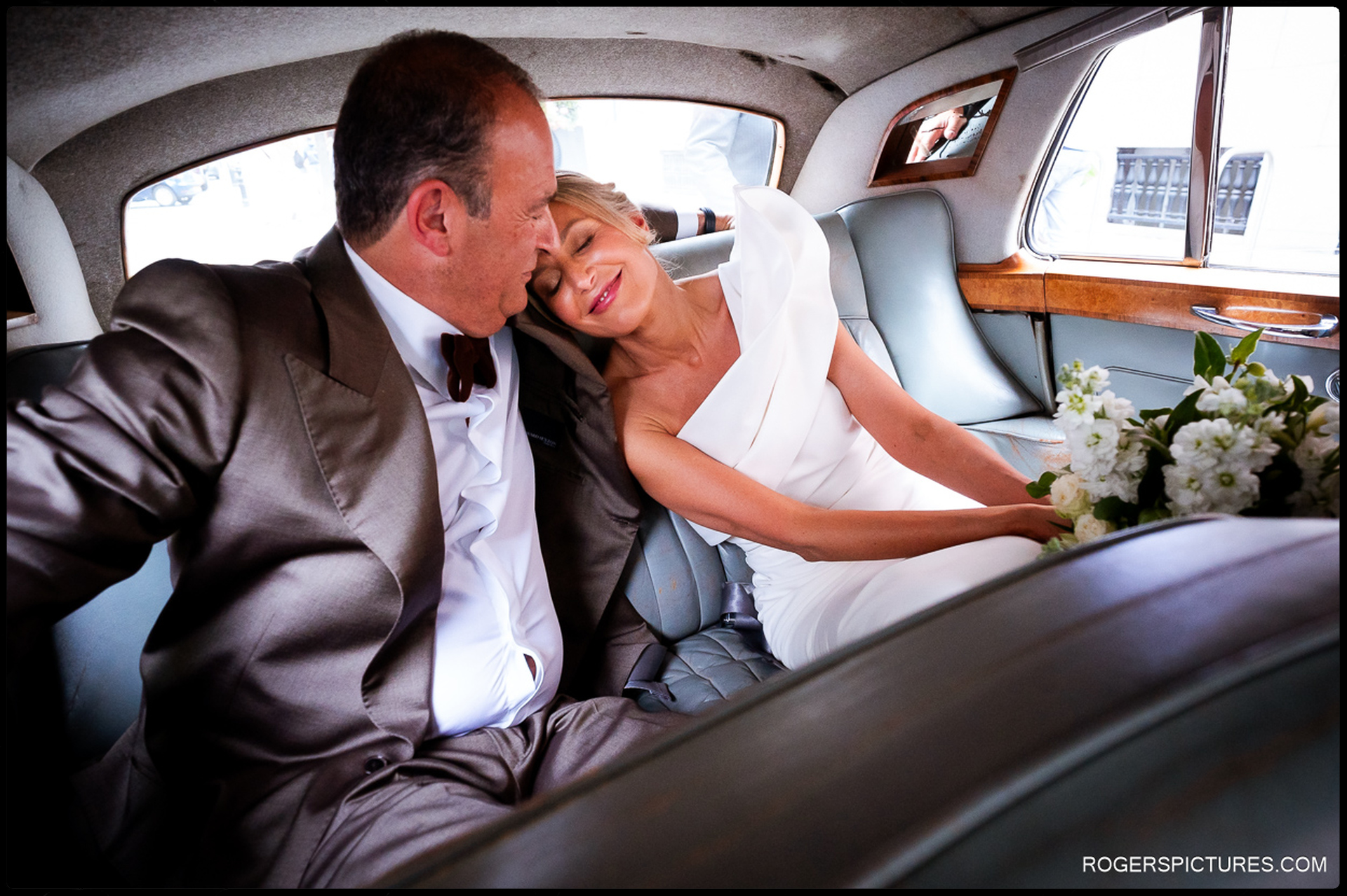 Bride and groom sitting together in the back of a vintage car, sharing a quiet moment as the bride leans her head on his shoulder.