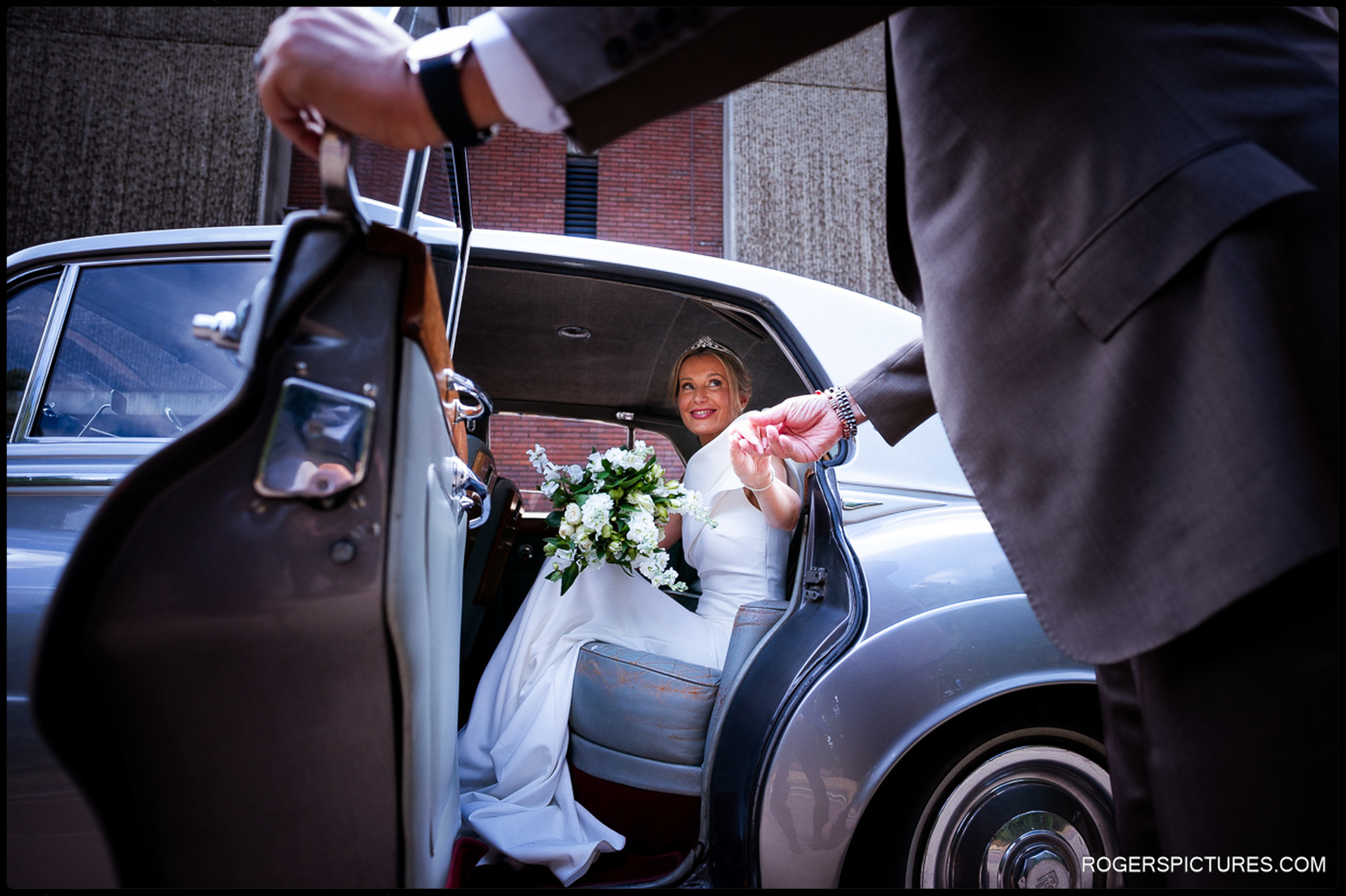 Bride smiling from the back seat of the vintage car, holding her bouquet as the groom opens the door and offers his hand.