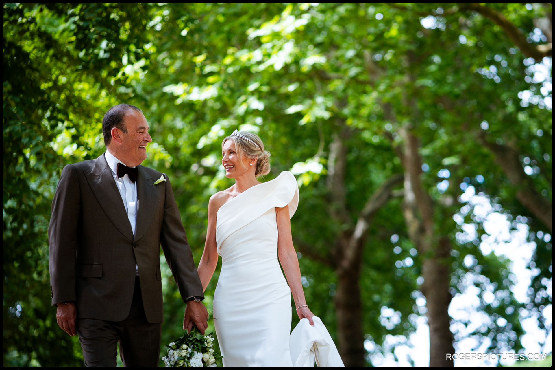 Bride and groom walking hand in hand through the green shade of Hyde Park, laughing together in the soft sunlight.