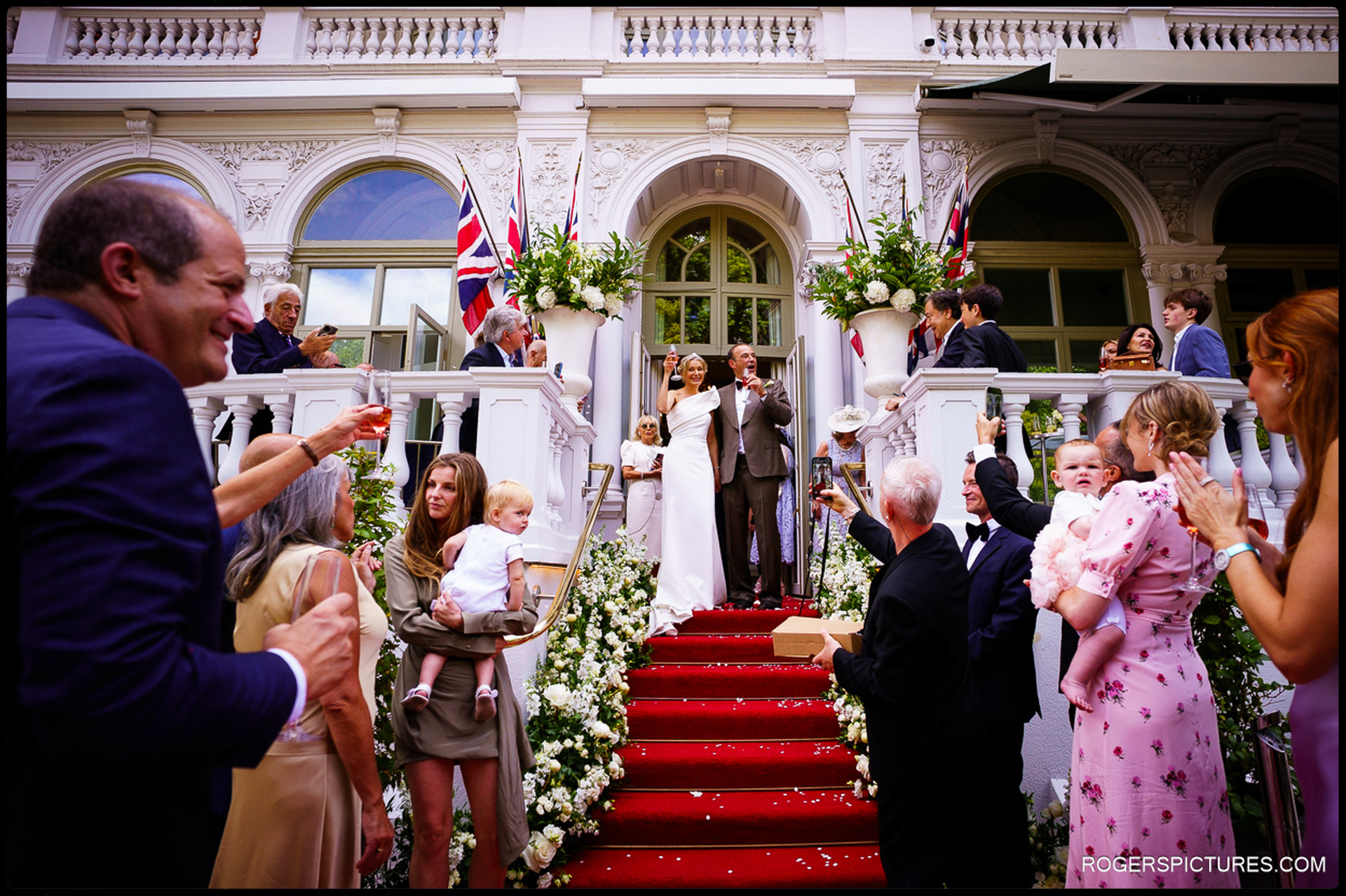 Bride and groom arriving at the Mandarin Oriental, greeted by guests gathered on the grand red-carpeted steps decorated with white flowers and Union Jack flags.