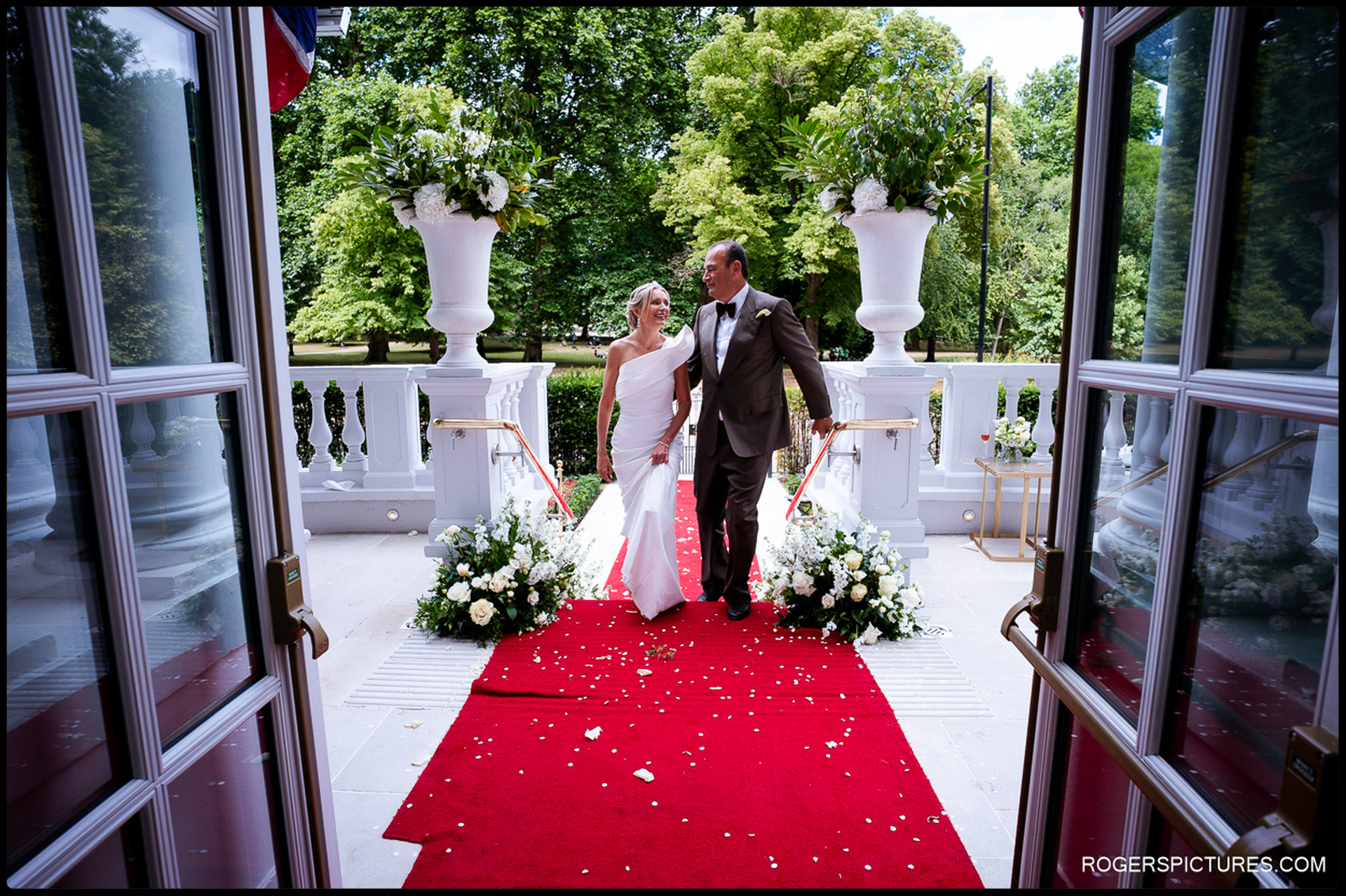 Bride and groom walking together down the red-carpeted terrace steps at the Mandarin Oriental, framed by large white floral arrangements and views of Hyde Park.