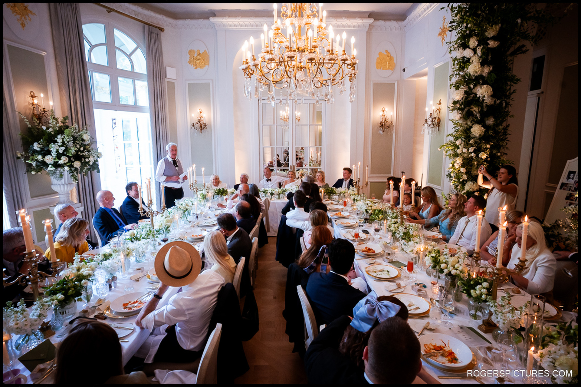 Wedding breakfast at the Mandarin Oriental with guests seated along a beautifully decorated table, listening to a speech under chandeliers and candlelight.