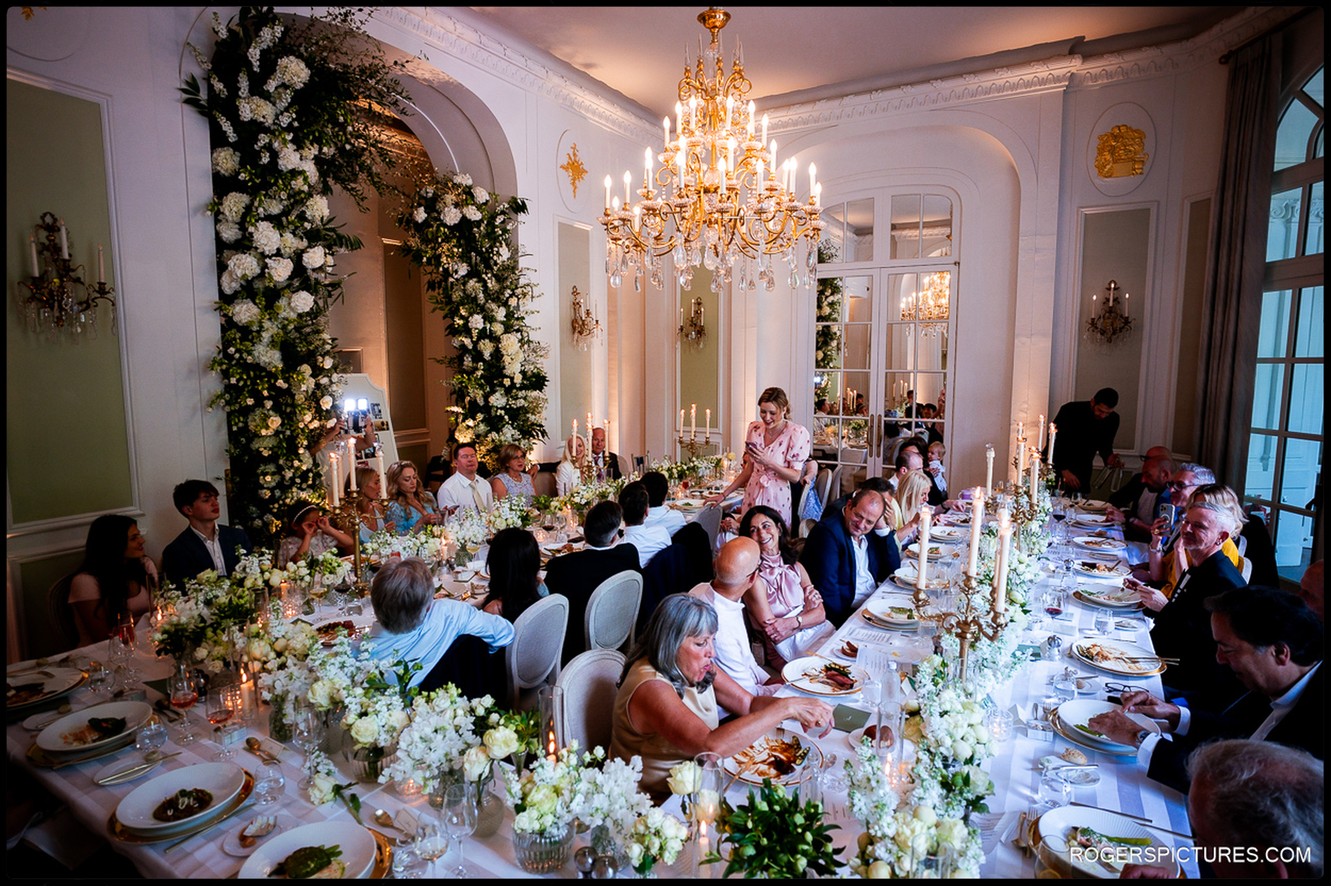 Guests enjoying speeches during the wedding meal at the Mandarin Oriental, surrounded by elegant white floral displays and golden chandeliers.