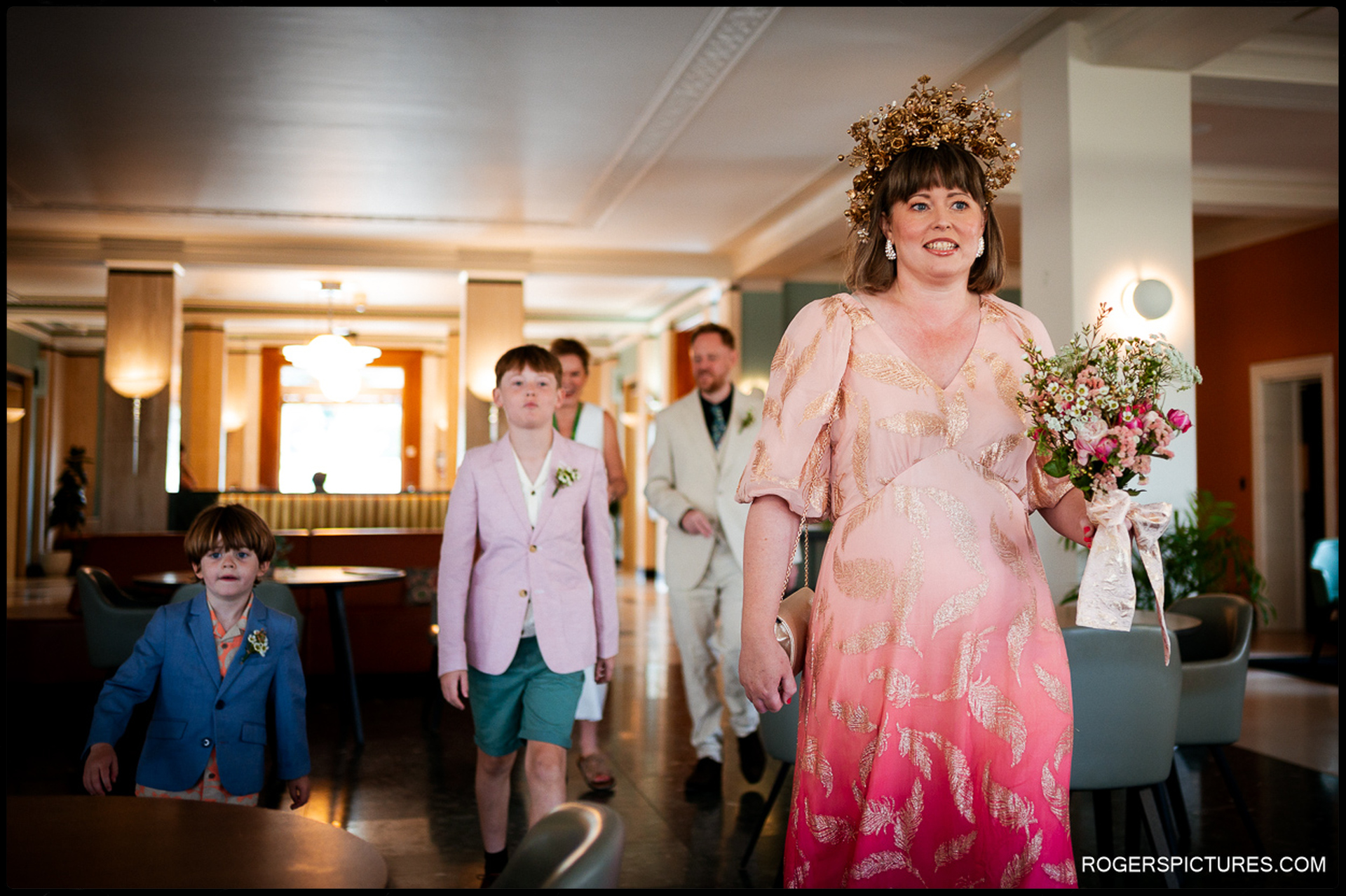 Bride arriving inside Waltham Forest Town Hall with her family, wearing a pink and gold dress and floral headpiece, walking with children towards the ceremony space.