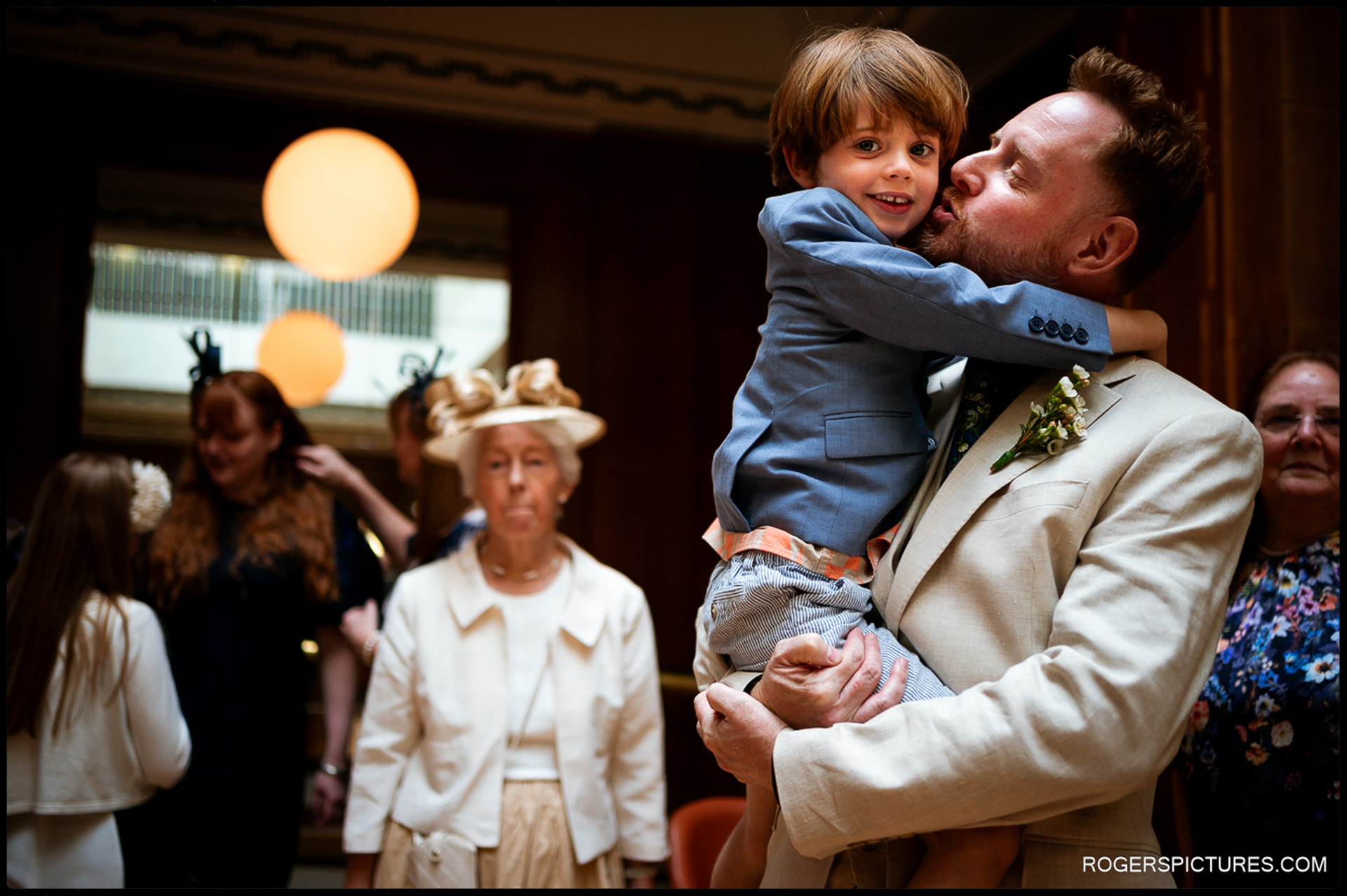 Groom holding and kissing a young child inside Waltham Forest Town Hall, surrounded by family before the wedding ceremony.