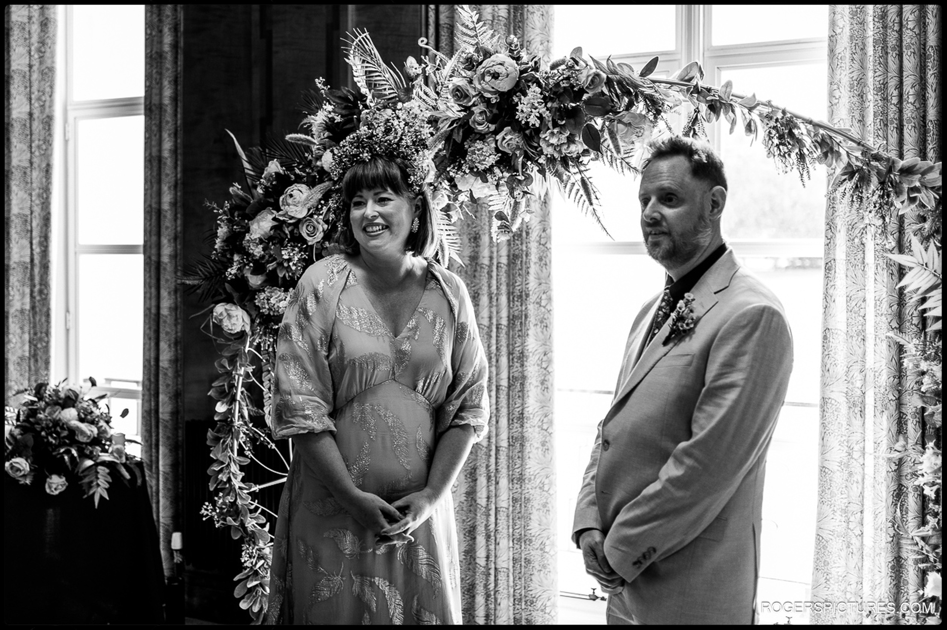 Bride and groom standing together under a floral ceremony arch inside Waltham Forest Town Hall, captured in a documentary black-and-white style.
