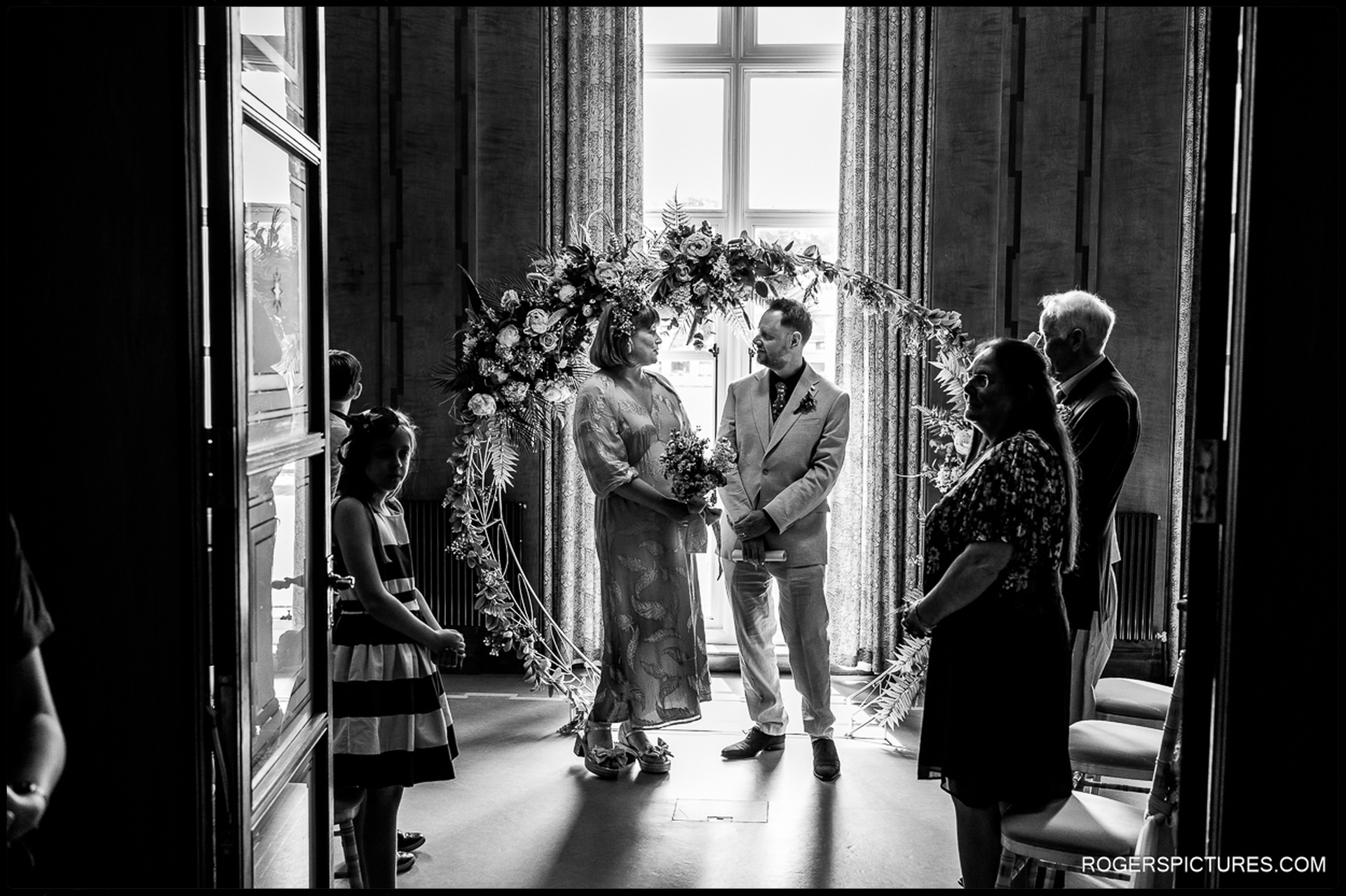 Bride and groom facing each other during their ceremony under a floral arch, with close family standing nearby, photographed from the doorway in black and white.