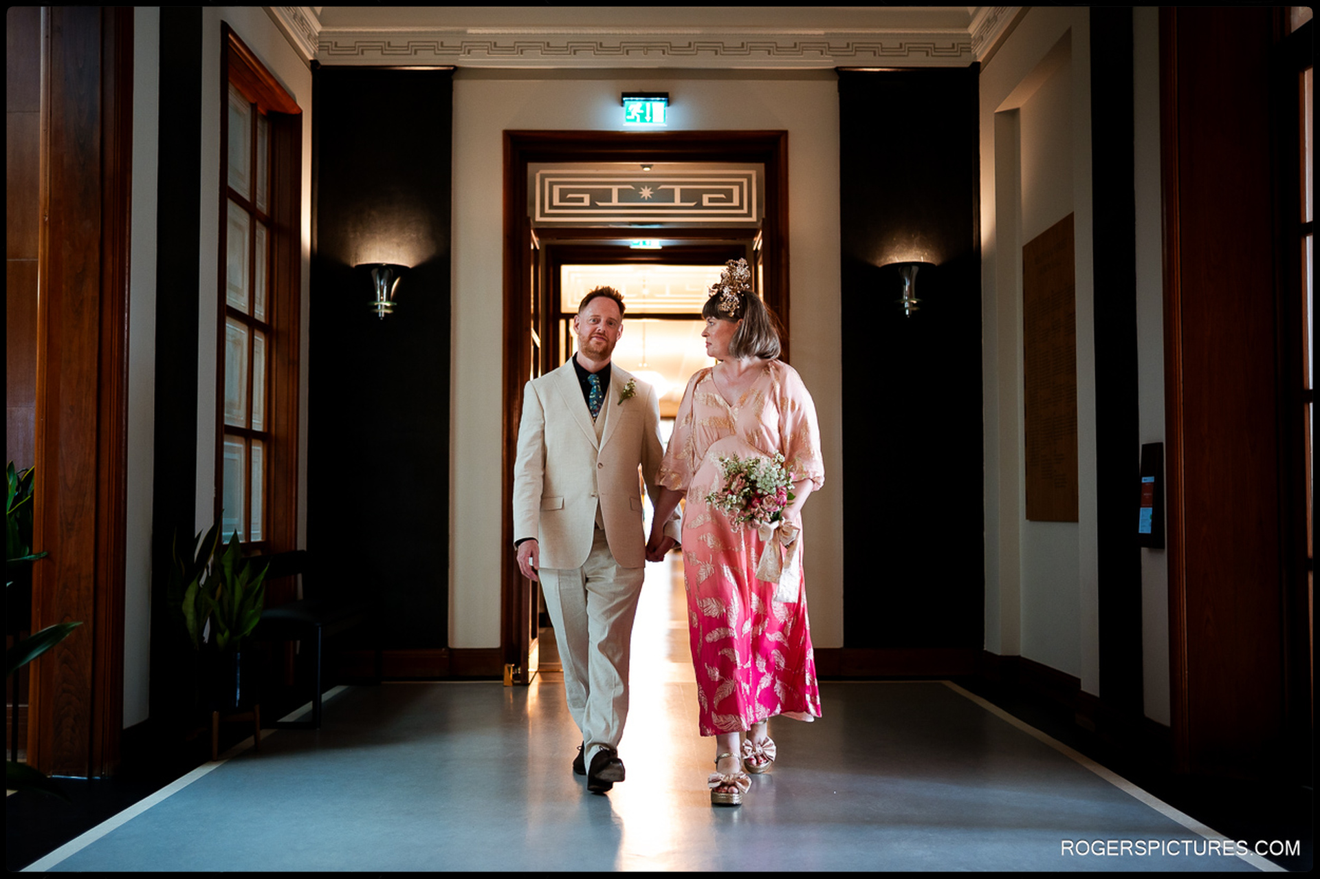 Couple walking hand-in-hand down a grand hallway inside Waltham Forest Town Hall after their ceremony, bathed in warm afternoon light.
