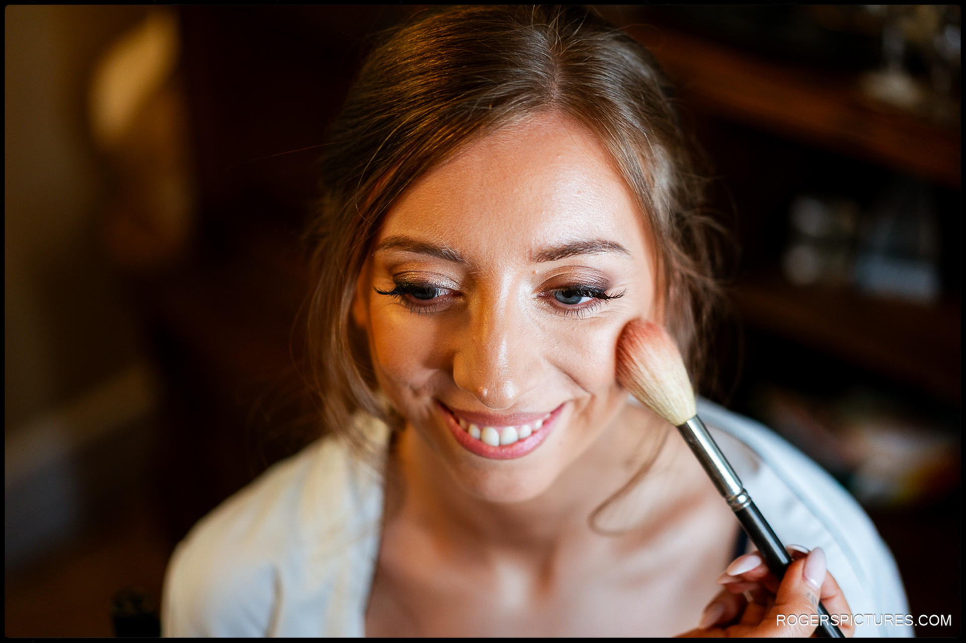 Bride smiling while having blush applied during morning preparations, soft warm light on her face.