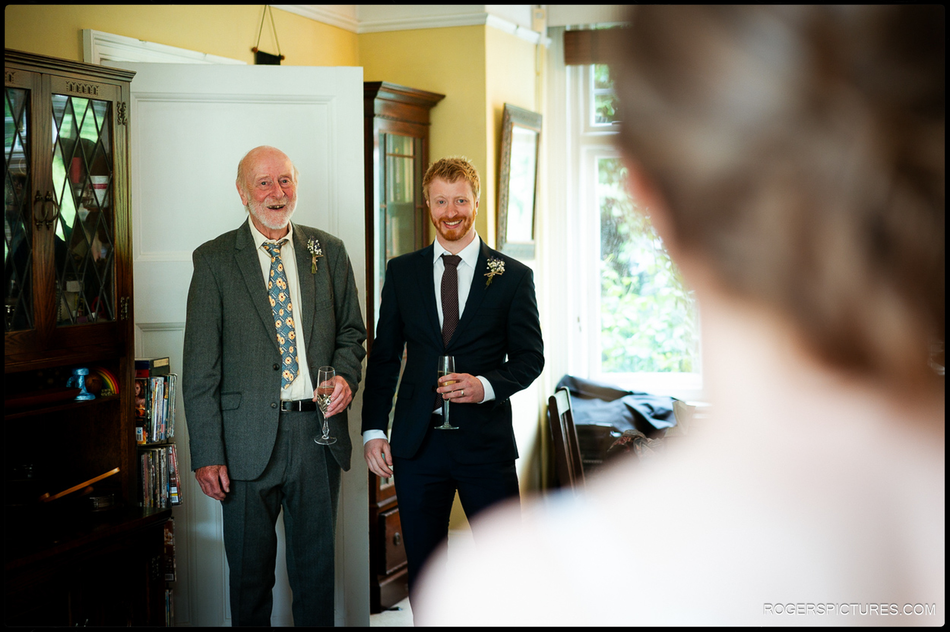 Groom and his father standing indoors smiling, holding champagne, reacting to seeing the bride.