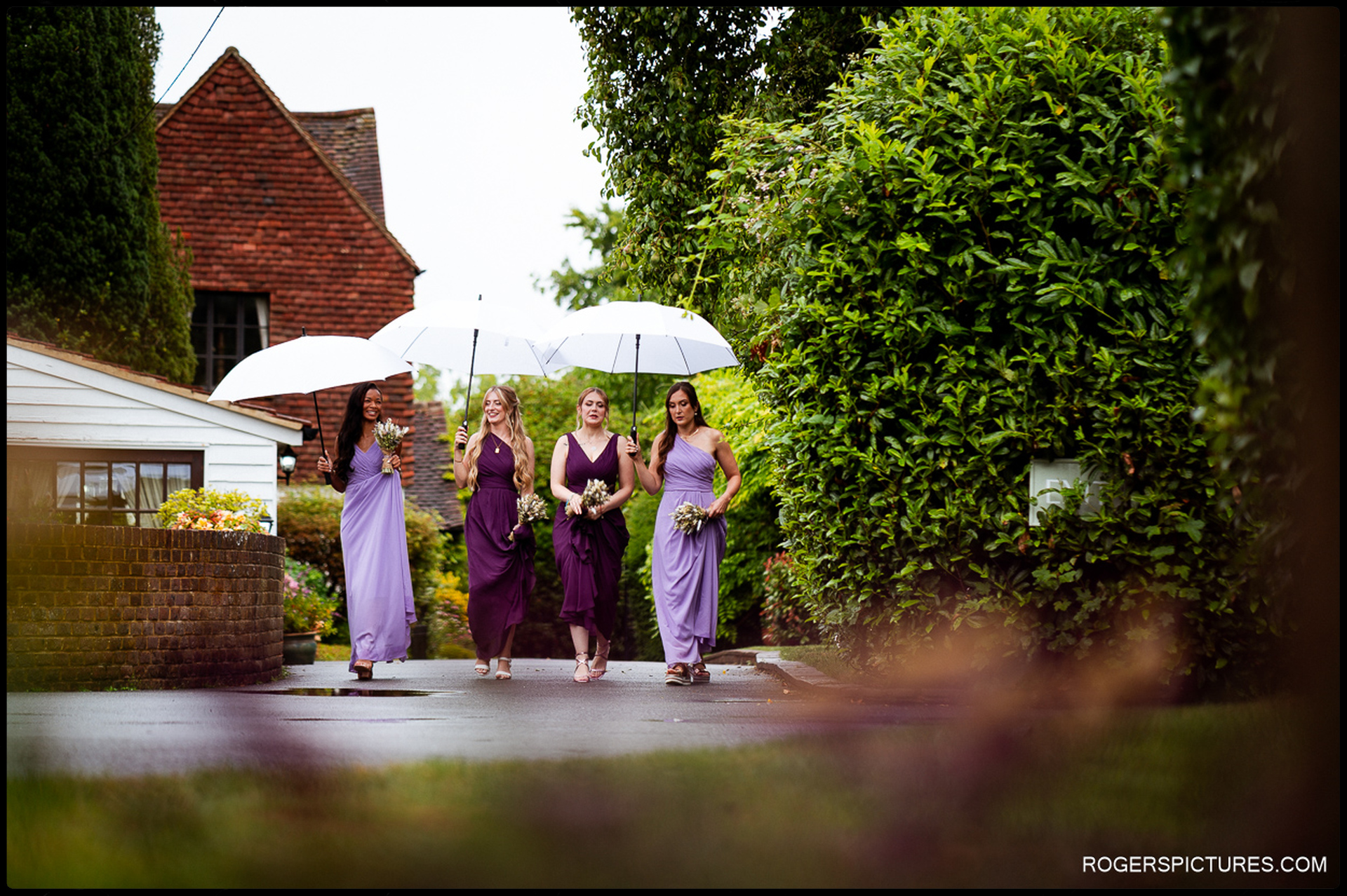 Bridesmaids walking down a garden path holding umbrellas and bouquets, approaching the venue.