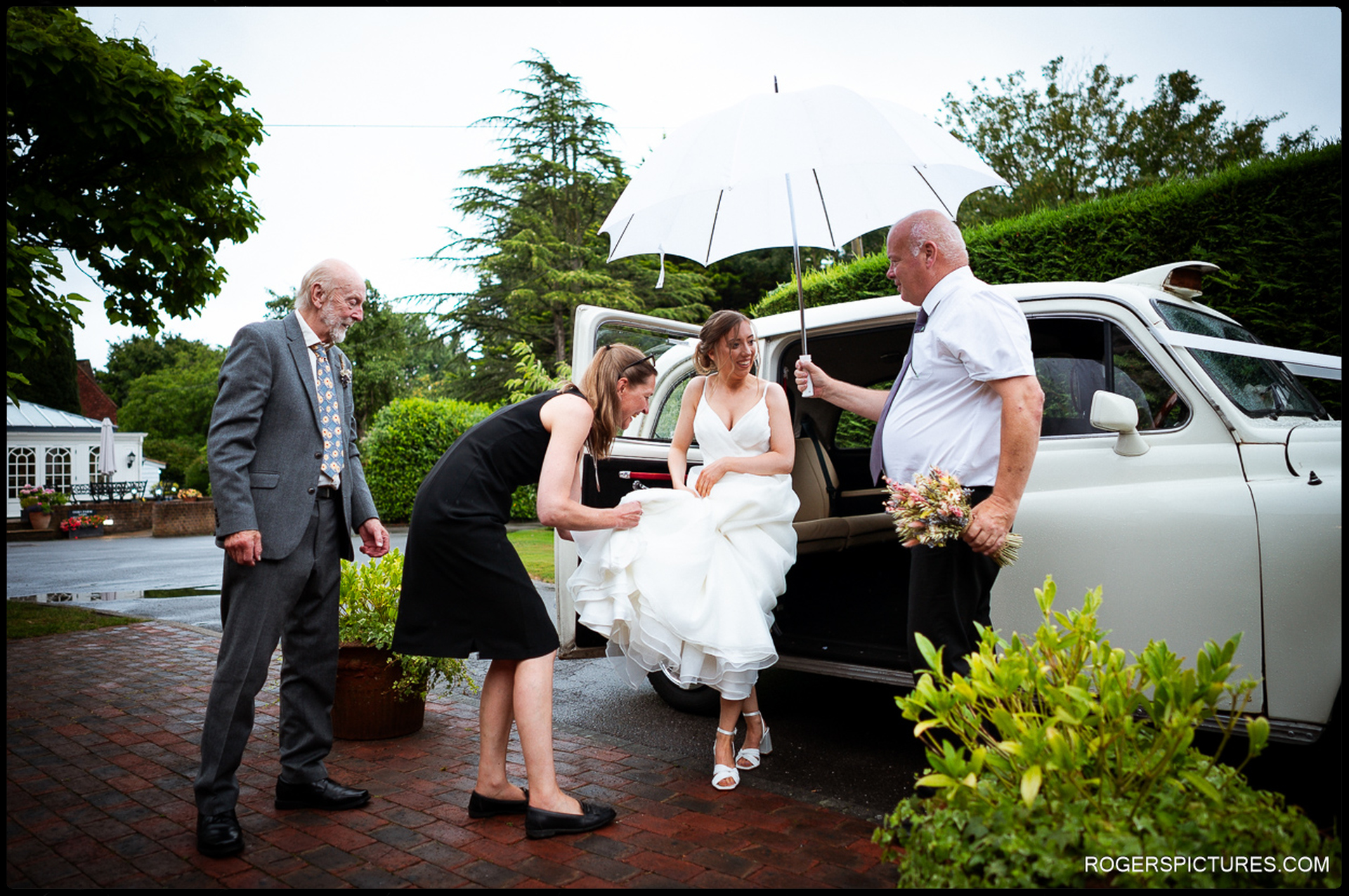 Bride stepping out of a classic wedding car in the rain, helped by family and the driver holding an umbrella.