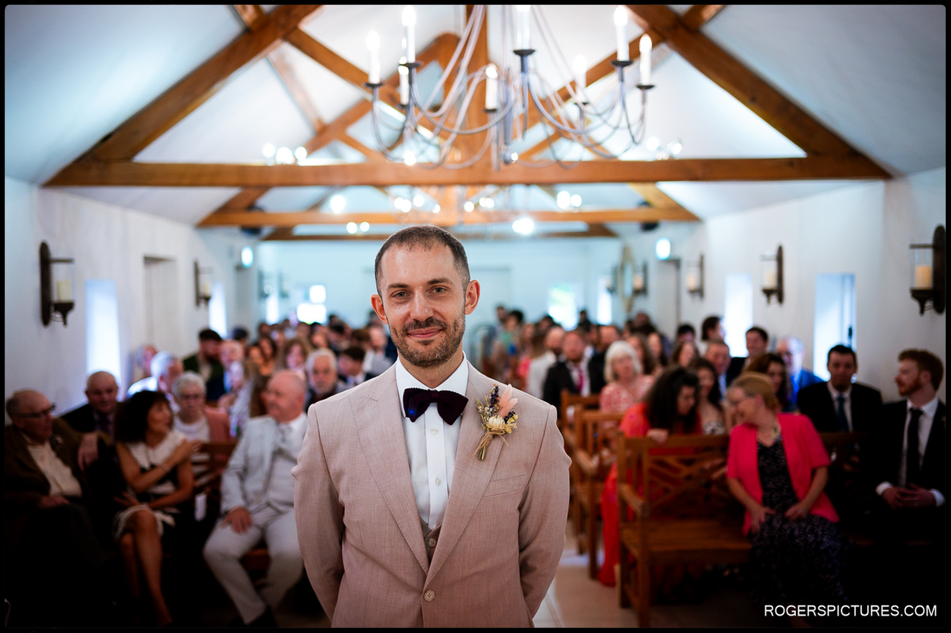 Groom standing at the front of the ceremony room looking toward the entrance with guests seated behind him.