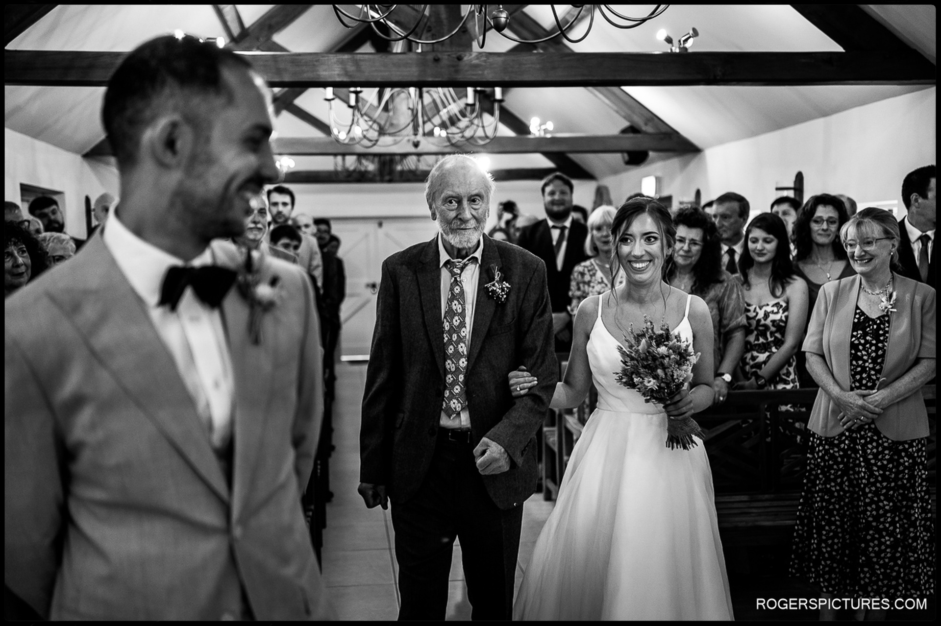 Bride walking down the aisle smiling, arm-in-arm with her father, guests watching in the background.