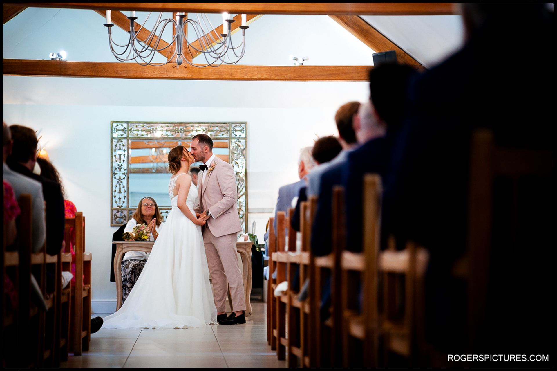 Bride and groom sharing first kiss at the ceremony table, hands held, guests seated on either side.